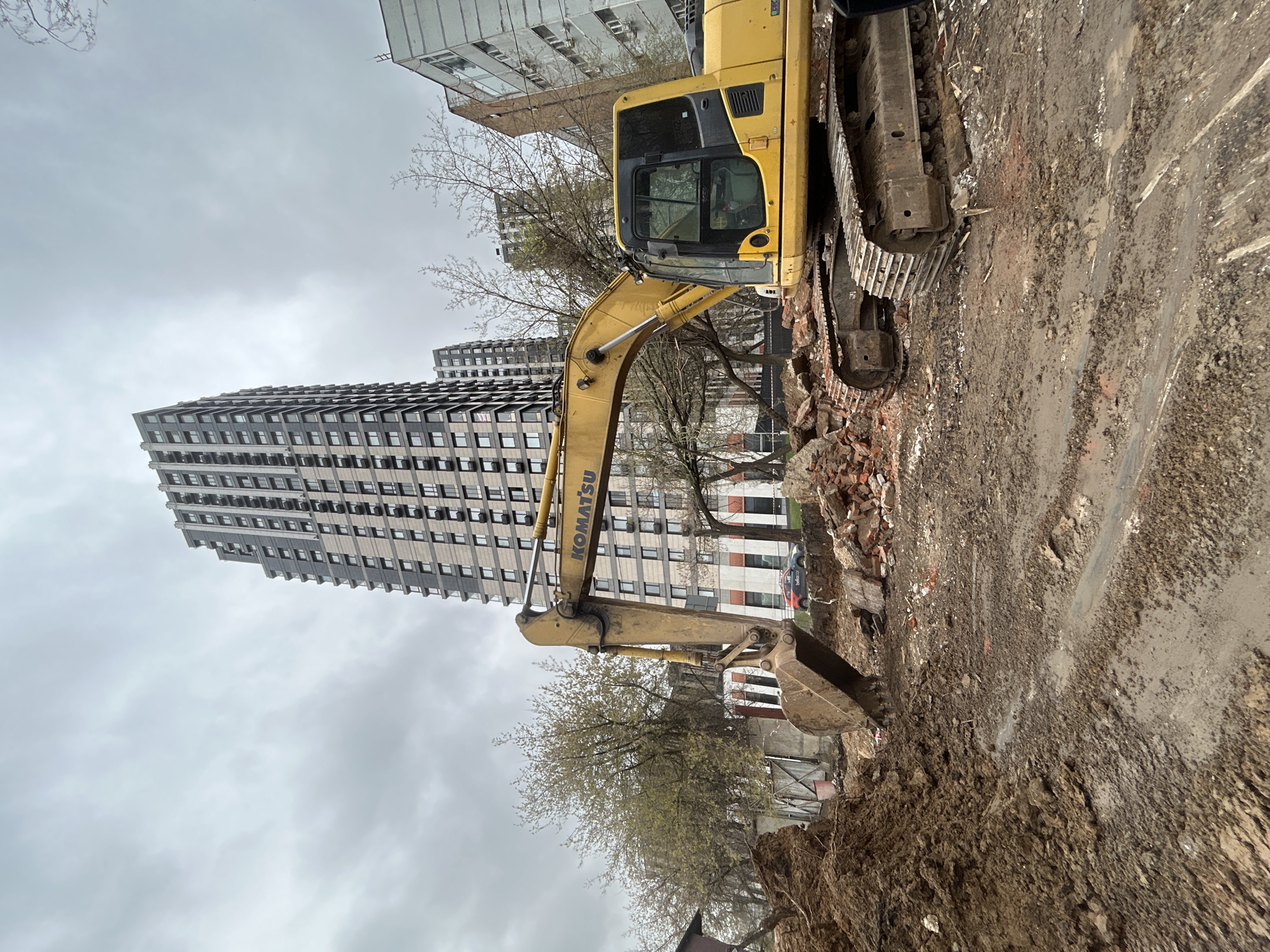 The image, presented in a portrait orientation and rotated 90 degrees clockwise, depicts an urban construction or demolition site in Moscow, Russia, under an overcast sky.
A large, yellow Komatsu tracked excavator is the central subject, prominently featured in the foreground and mid-ground. Its arm is extended forward, and the bucket is positioned near or in the deeply rutted and muddy ground. Significant construction debris, including broken bricks and rubble, is scattered around the excavator's tracks and bucket, indicating active demolition or site clearance. The text "KOMATSU" is clearly visible on the excavator's arm.
In the background, several tall, modern residential buildings rise against the grey, cloudy sky. A distinctive dark, multi-story building on the left side of the frame features a stepped or zig-zag architectural facade with numerous windows. Further to the right, another building, possibly older, with visible air conditioning units, is partially observed. Sparse trees, either bare or just beginning to bud, are interspersed between the foreground machinery and the distant buildings. A partially obscured car, likely a service vehicle, is visible in the background, with "bellco.ru" printed on its side. The overall scene conveys a sense of ongoing urban development, renovation, or demolition, with the cloudy weather casting a subdued light over the industrial setting.