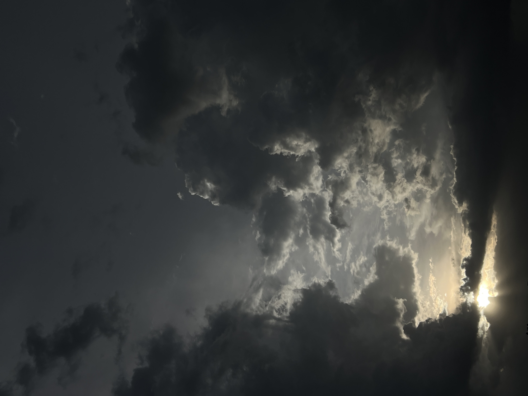 The image captures a dramatic sky filled with dark storm clouds, possibly indicative of impending rain. A patch of brighter, white-gray clouds is visible near the center, through which sunlight is breaking, creating a contrast between light and shadow. The location is not ascertainable from the image, and the city of Gura Top, Nigeria cannot be seen within the frame. There are no visible people, subjects, objects, actions, or interactions. The environment depicted is solely the sky.