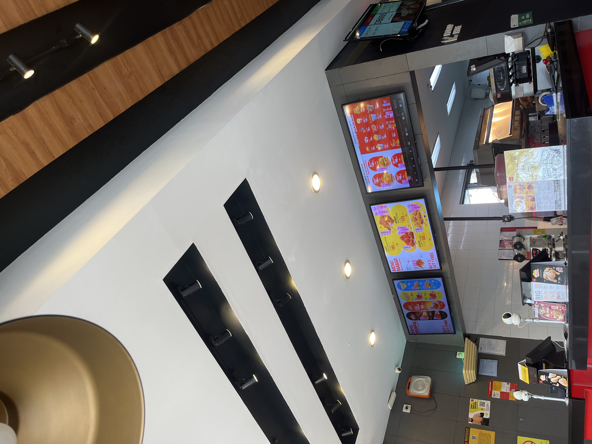 The image shows the interior of a fast-food restaurant, likely a chain specializing in fried chicken. The scene is viewed from a slightly elevated perspective, looking towards a counter area.
Dominating the upper portion of the image are two parallel black rectangular structures, likely ceiling elements, with embedded spotlights. Above these, a textured wooden ceiling is visible, angled and interspersed with black panels, suggesting modern architecture. A large, golden lampshade hangs from the ceiling on the left.
Below the ceiling structures, a series of digital menu boards are displayed on the wall. The menus feature images of food items, including chicken, burgers, and fries, along with prices in Indonesian Rupiah. One menu board prominently displays "MENU NAMPOL" and offers a "BUY 3 GET 1 FREE" promotion. Another board shows "MABAR Fire Chicken" for Rp 173,000.
In the foreground, a black counter extends across the width of the image. On the counter, various items are visible, including a touch screen monitor for ordering, receipt printers, a tape dispenser, and what appears to be a cash register system. Several menus or promotional flyers are placed on the counter, some with images of fried chicken. A small security camera is mounted on the wall above the counter.
The walls are a mix of grey tiled surfaces and dark grey panels. Yellow and white signs with Indonesian text are affixed to the walls. A beige shelving unit is mounted above the menu boards.
While no specific individuals are clearly depicted, the presence of the counter, ordering systems, and menus indicates a service area within a restaurant environment. The overall impression is of a clean, well-lit, and modern fast-food establishment. The text on the menus and promotional materials, such as "MABAR" and prices in Rupiah, strongly suggests the location is in Indonesia, specifically Tangerang, given the prompt's context.