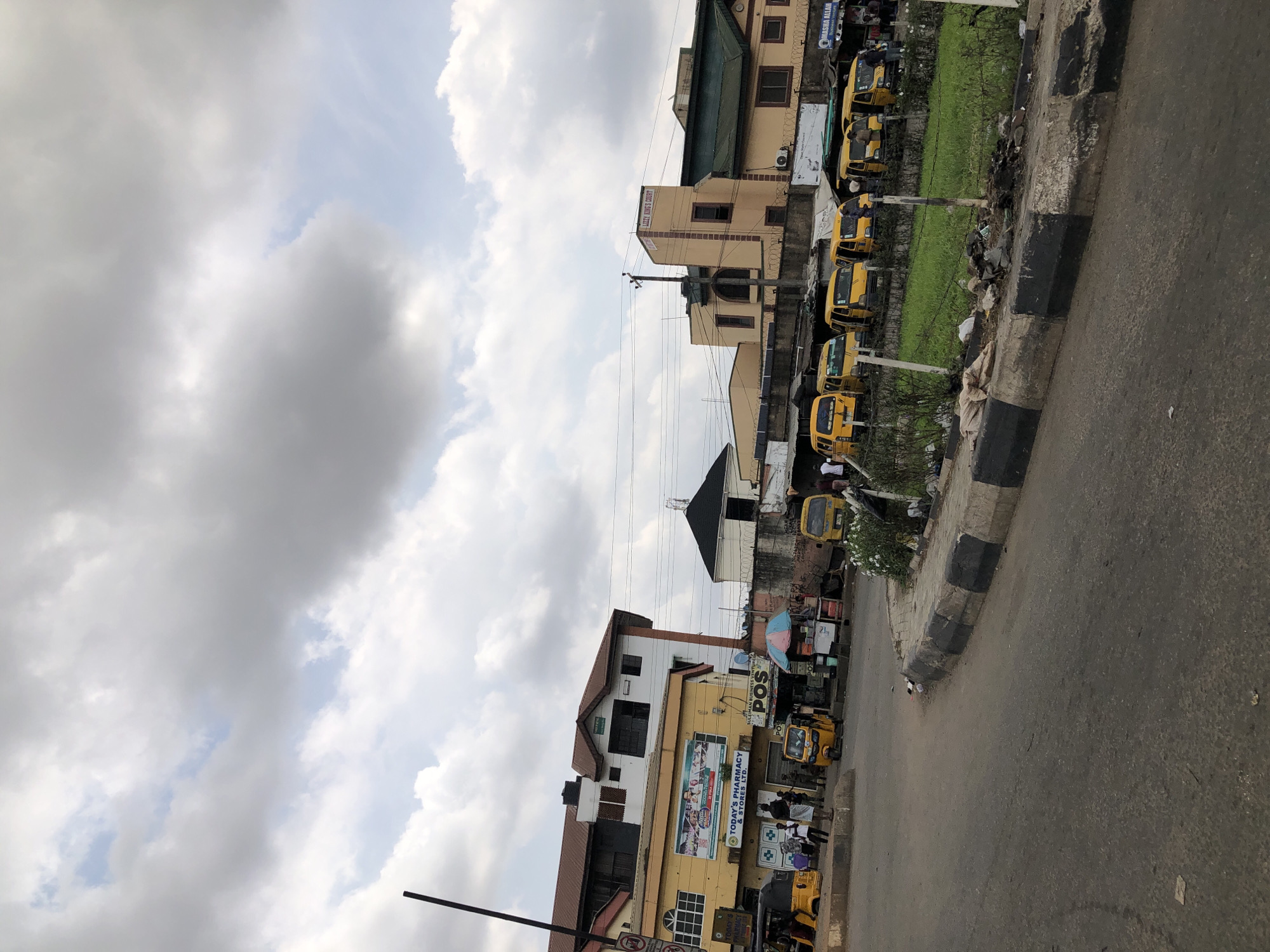 The scene is a street view in Isheri, Nigeria. The sky is cloudy. On the right side of the frame, a row of yellow commercial tricycles (often called "Keke Napep") is parked along the curb, next to a small patch of green space bordered by a painted curb. On the left, there are commercial buildings, including a pharmacy with a large sign reading "TODAY'S PHARMACY & STORES LTD.", a POS business, and a building named "LIZZY KING'S COURT". People can be seen standing and walking near the pharmacy and other businesses. A black pole stands on the left side of the frame. The street surface is visible.