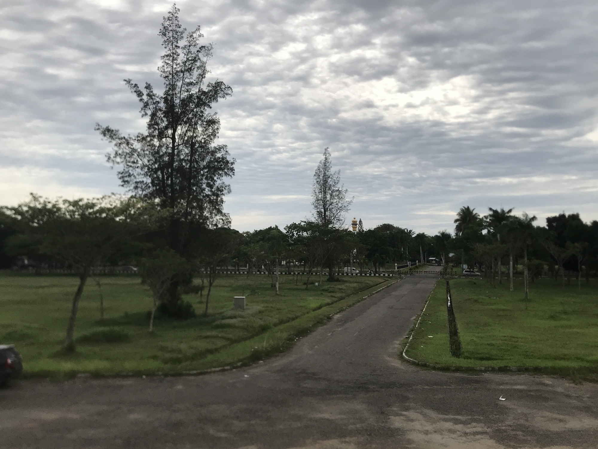 The image shows a paved road in Tambontunong, Indonesia, surrounded by grassy fields and various trees under a cloudy sky. In the distance, there is a building with a gold-colored dome. A car is partially visible on the left side of the image. The road curves into the distance, lined with trees and landscaping.