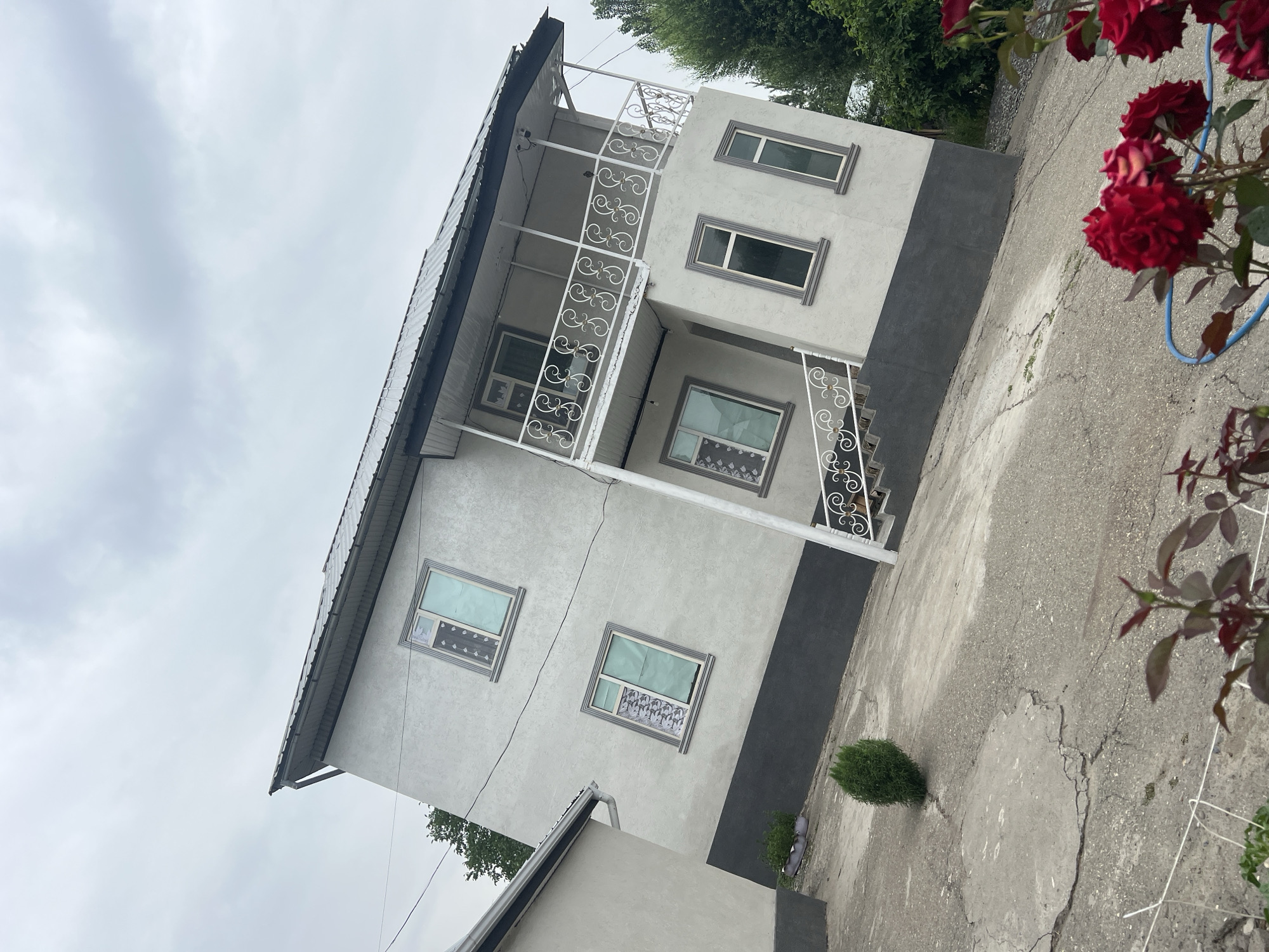 The image captures a two-story house in the city of Kara-Dıykan, Kyrgyzstan. The house has a light grey stucco exterior with a dark grey foundation. It features a metal roof and several windows with decorative frames. A white balcony with an ornate railing is present on the upper floor, accessible by an external staircase. The surrounding environment includes a cracked concrete driveway, a small green bush, and lush green trees in the background. Red roses are visible in the foreground, with a blue hose coiled nearby. The sky is overcast with grey clouds. The perspective of the image is tilted, suggesting it was taken from a low angle. No people are visible in the scene.