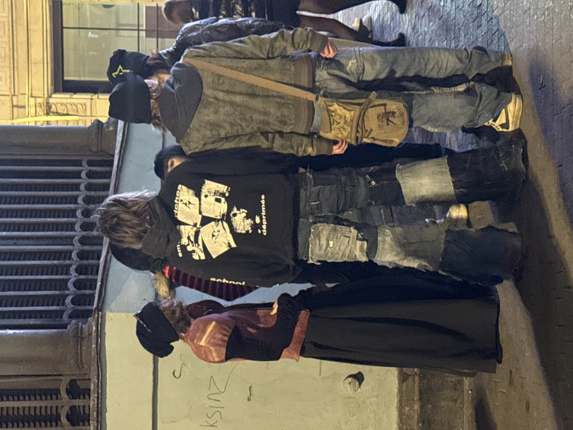 A group of people stand on a sidewalk in Saint Petersburg, Russia. The image is taken at night, with artificial light illuminating the scene. The group comprises several individuals with diverse clothing styles. One person wears a black hoodie featuring white graphic images and the word "deprimes." Another person wears a hooded jacket, light blue jeans, and a crossbody bag. A third person is wearing a long black skirt and a dark red top. The sidewalk is concrete and has some graffiti on it. A metal grate is visible on the left side of the image. The background shows part of a building with illuminated windows.