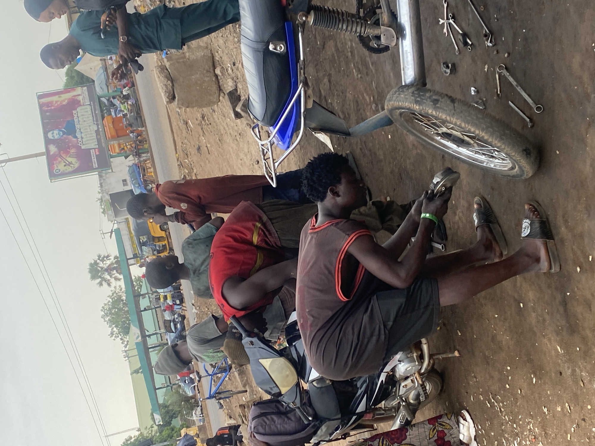 The image depicts a motorcycle repair scene in Sabon Tasha, Nigeria. Several men are gathered around motorcycles, with one man seated on a motorcycle, seemingly working on its components. Various tools are scattered on the ground, indicating ongoing repair activities. In the background, there is a street scene with buildings and advertisements, suggesting an urban environment. Other individuals are standing and observing the repair process.
