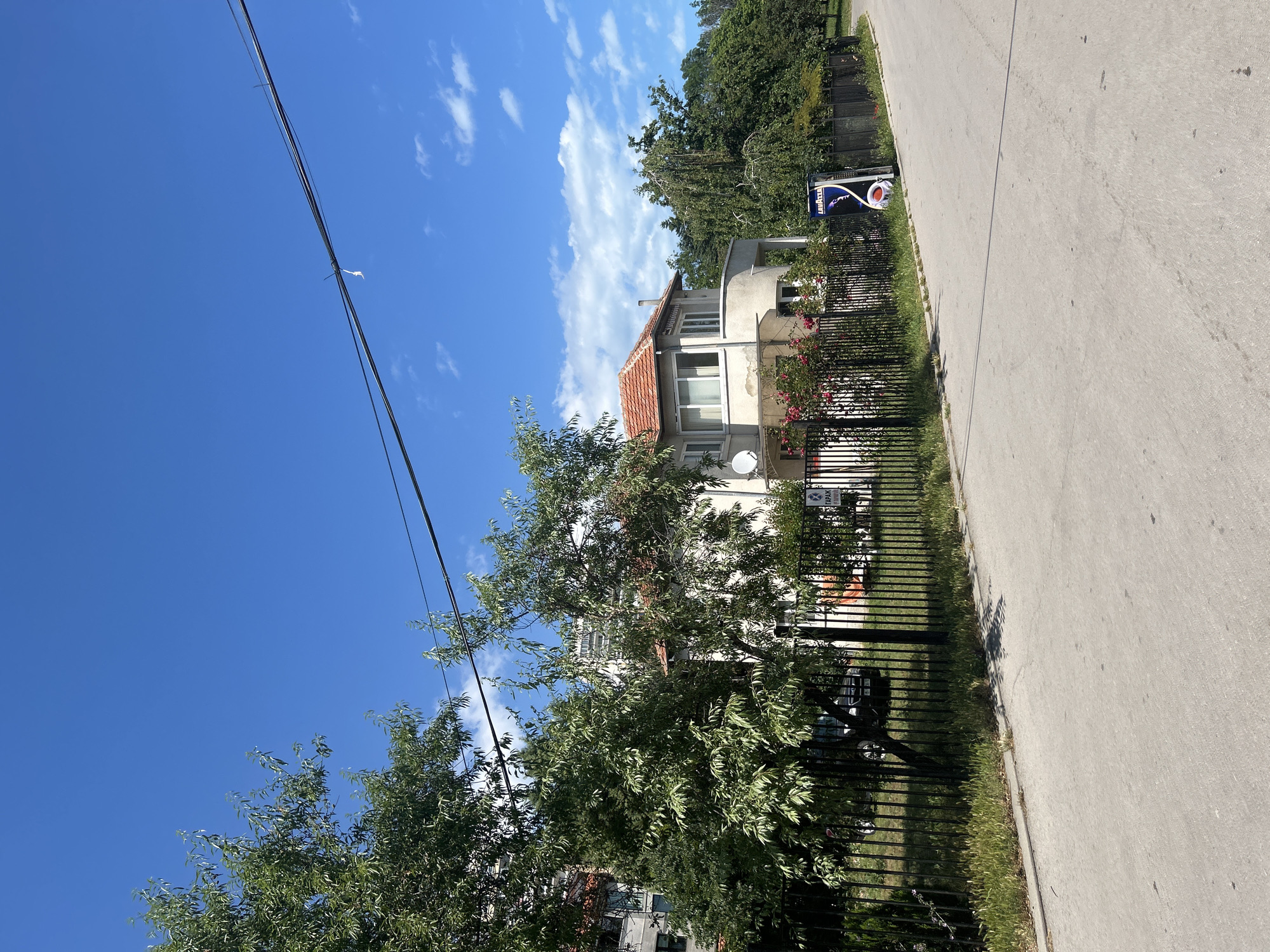 The image depicts a street scene in the city of Varna, Bulgaria. In the foreground, a paved road stretches towards the right. A black metal fence borders the left side of the road, behind which is a green lawn and trees. A two-story house with a red tile roof is visible behind the fence, featuring a curved balcony on the second floor. A satellite dish is mounted on the side of the house. Further back, more trees and buildings are visible, including a taller, modern apartment building. The sky is a clear blue with scattered white clouds. A street sign with Cyrillic text that appears to be "ГАРАЖ" (Garage) is visible near the fence on the right side, along with a blue and white advertisement for Lavazza coffee. The overall scene suggests a residential area under a bright, sunny sky.