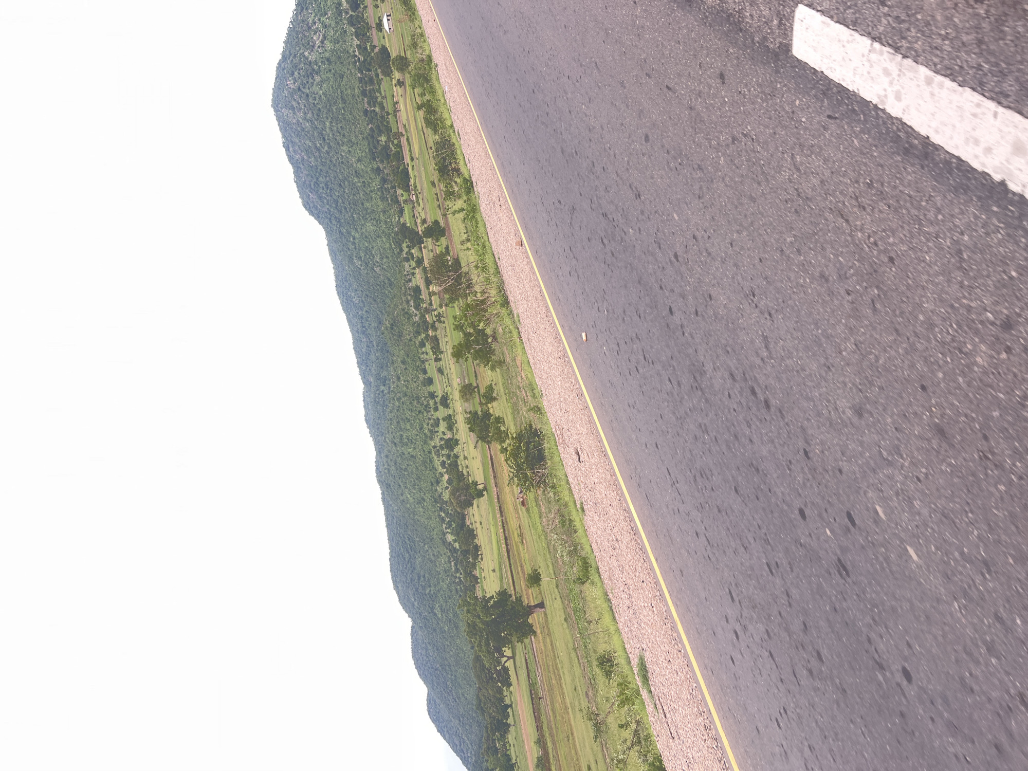 The image shows a rural landscape with a paved road in the foreground. The road has a yellow line marking its edge. To the left of the road is a gravel shoulder with sparse vegetation. Beyond the shoulder, a green field stretches towards a series of rolling hills covered in trees and vegetation. The sky is bright and overcast. A small white building with a dark roof is visible in the distance on the hillside. The scene suggests a natural environment, possibly outside of a built-up area.