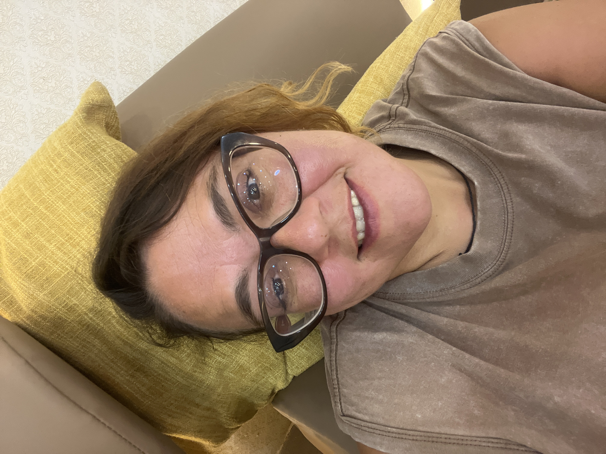 A woman wearing glasses and a brown t-shirt lies on a yellow couch, looking at the camera. The couch is next to a wall with textured wallpaper. The scene is indoors.