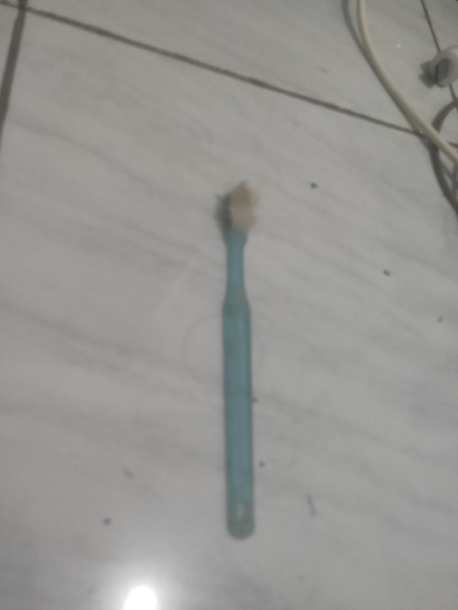 A single, light blue toothbrush lies on a light-colored, tiled floor. The toothbrush is oriented vertically and appears to be slightly worn, with some light-colored residue on its bristles. The tiled floor has visible grout lines, creating a grid pattern. A thin, light-colored cord, possibly from an electrical appliance, is visible in the upper right corner of the image. The lighting in the scene creates a bright, circular reflection on the floor in the lower portion of the image. The background is out of focus. The scene is indoors, consistent with a bathroom or similar tiled area. There are no people present.