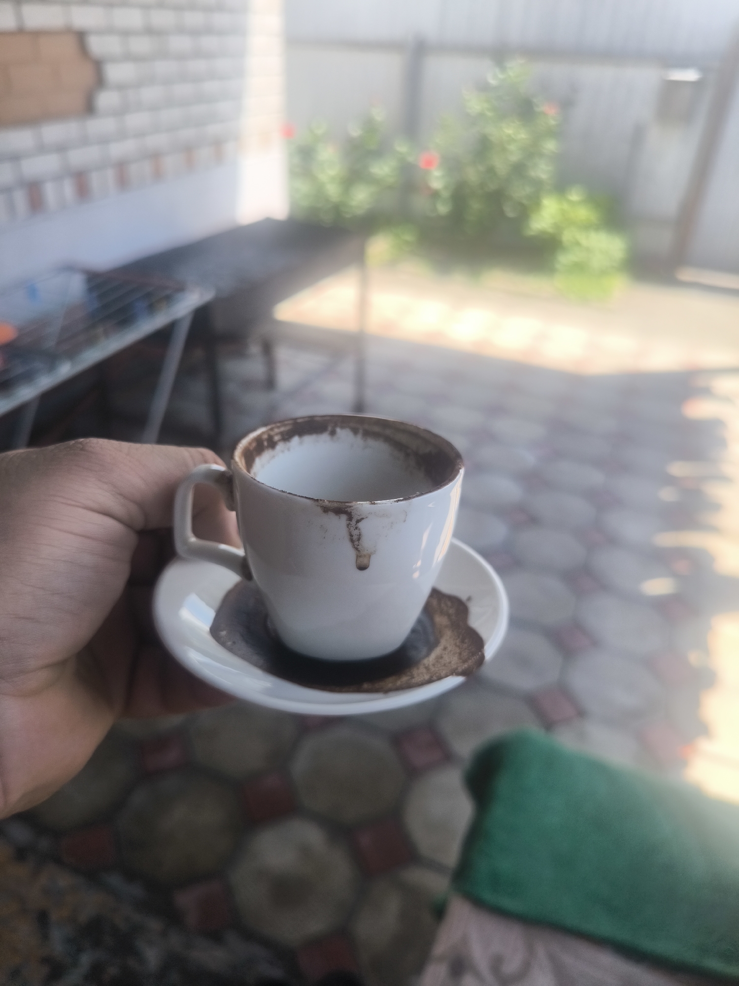 A human hand, likely an adult's, holds a white ceramic coffee cup with a matching saucer, observed from a slightly elevated, close-up perspective. The cup and saucer are heavily stained with dried dark brown coffee grounds, with noticeable drips on the exterior of the cup and a thick residue accumulated on the saucer. The hand, positioned in the lower-left quadrant of the frame, is the primary subject in focus.
The background is significantly blurred. To the immediate left, a light-colored brick wall with a reddish-brown damaged section is visible, beneath which a dark, oblong surface and metallic rack-like structures are present. Further back and to the right, green foliage with indistinct red spots, presumably flowers, indicates a garden area. A light-colored fence or wall defines the distant perimeter. The ground beneath the cup and extending to the right features a pattern of blurred hexagonal or octagonal tiles in various shades, with faint reddish accents. A segment of dark green fabric, possibly a towel or blanket, lies in the lower-right foreground, partially obscuring another patterned fabric surface. The scene is illuminated by natural daylight, casting subtle shadows and highlights. The location is an outdoor patio or veranda in the city of Unknown, Unknown.