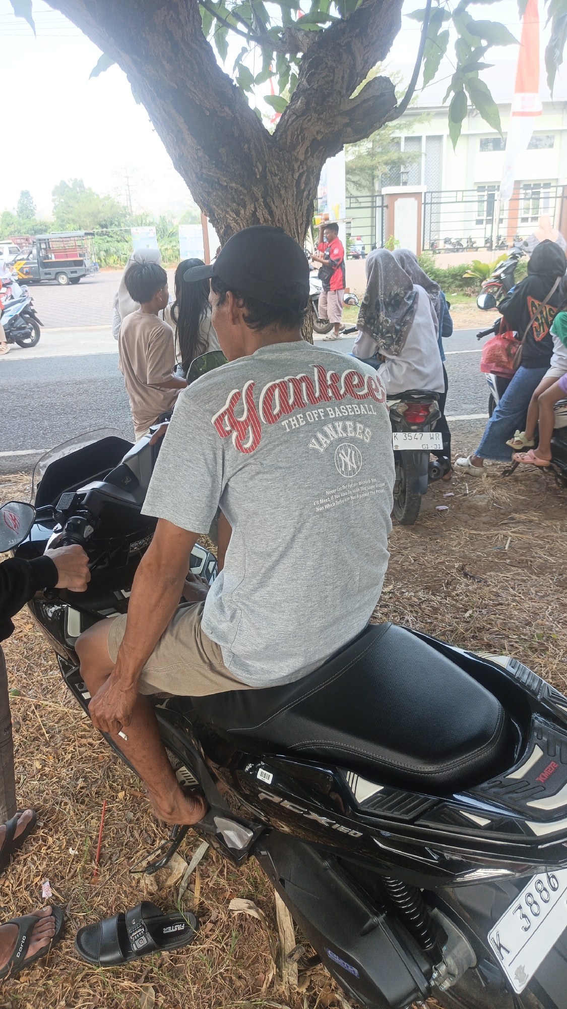 In an outdoor, possibly urban setting, a man wearing a black baseball cap and a grey t-shirt with "Yankees" and baseball imagery on it, is seated on a black motorcycle. He is wearing tan shorts and has a cigarette in his hand. The motorcycle is parked on a dirt and dry grass area, with some foliage visible. In the background, there are other people, some on motorcycles, and a building. There is also a tree with prominent branches in the foreground. The sky appears bright, suggesting daytime. The scene is captured from a medium-low angle.