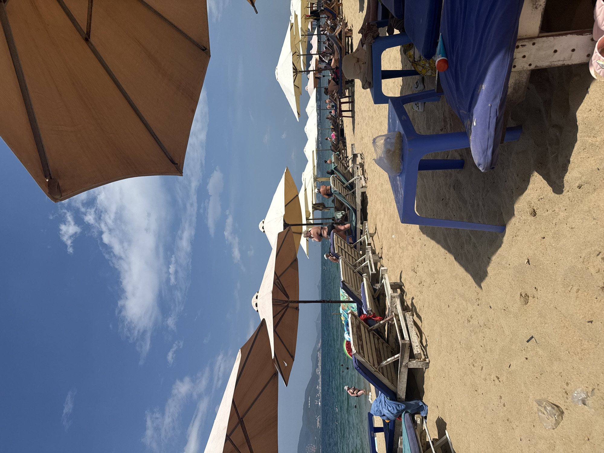 This is a daytime, eye-level, full shot of a crowded beach in Nha Trang, Vietnam. The sand is light brown, with several rows of empty and occupied lounge chairs. Large, tan umbrellas provide shade over some of the chairs. The ocean is visible in the background, with a distant coastline and mountains under a partly cloudy blue sky. Several people are in the water, and others are relaxing on the beach. On the right side of the frame, a blue plastic table holds a bag of snacks and a tube of sunscreen. The scene is typical of a popular tourist beach.