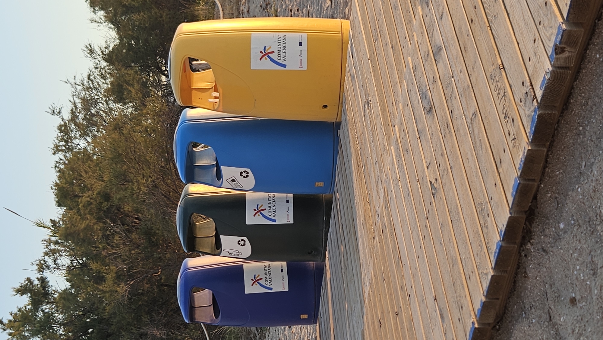 This media file shows a row of four colorful recycling bins on a wooden boardwalk. The bins are stacked vertically, with a yellow bin on top, followed by blue, dark green, and another blue bin at the bottom. Each bin has an opening for waste disposal. Some of the bins have stickers with a logo that reads "COMUNITAT VALENCIANA" and some recycling symbols.
The scene is set outdoors, with trees and vegetation in the background. The ground appears to be sandy, consistent with a beach or park setting. The wooden boardwalk is made of planks and runs diagonally through the frame, suggesting it might lead to a beach or other area.
The lighting suggests it is either early morning or late afternoon, with soft sunlight casting shadows and highlighting the texture of the wood. The sky is clear and pale, indicating fair weather. There are no people visible in the image. The overall impression is one of an organized public space, likely a beach area given the location context of Deveses, Spain. The bins are prominently displayed objects, representing a focus on waste management and environmental consciousness in the setting.