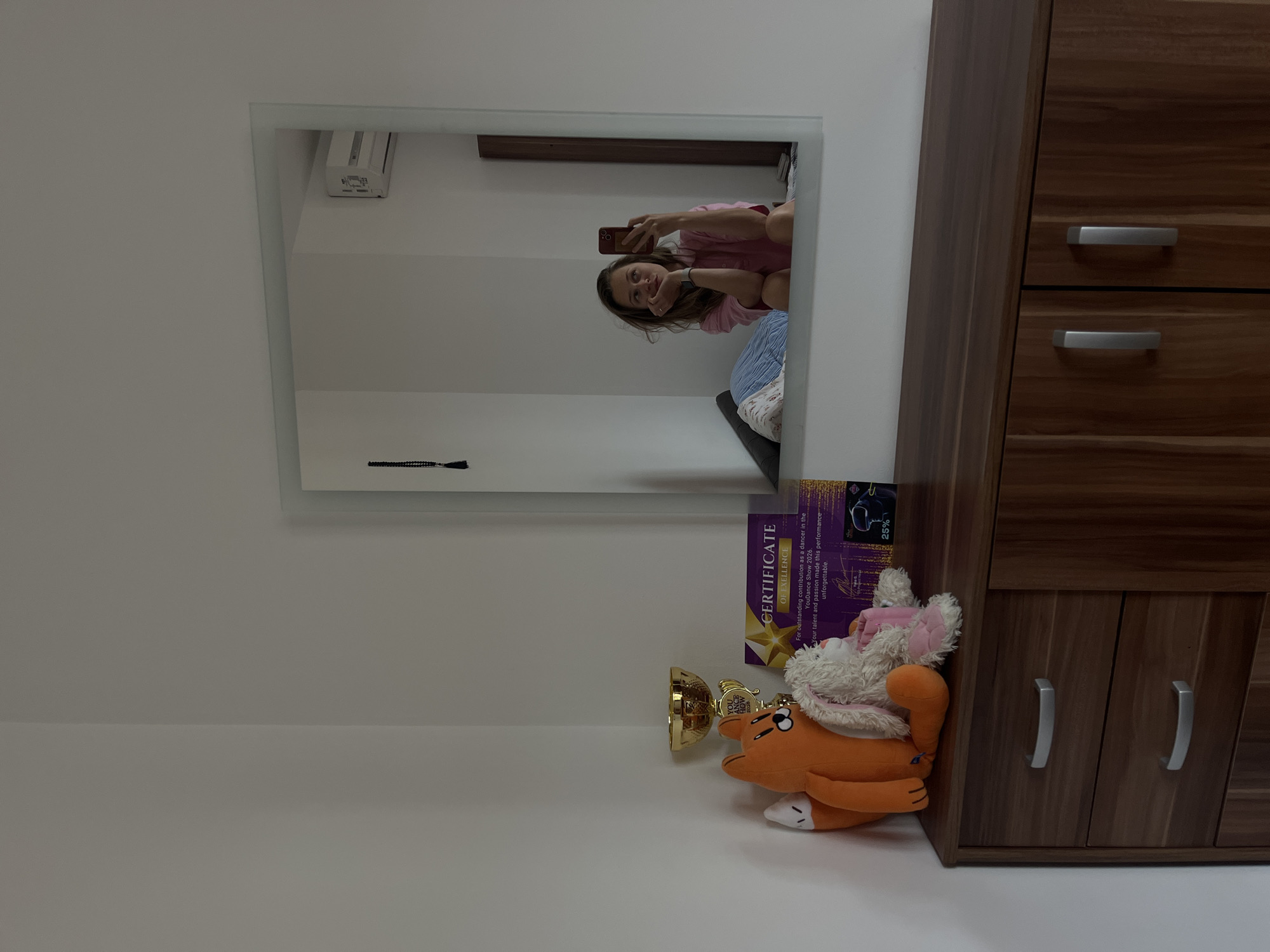 The photograph shows an interior setting. On the right of the frame, there is a wooden cabinet with multiple drawers and metal handles. To the left, a mirror reflects a woman taking a selfie with her phone. The woman is wearing a pink top and appears to be sitting on a bed. In front of the cabinet, on a white surface, there are a few items: a trophy, a stuffed animal resembling an orange dog, and a certificate of excellence. The certificate contains the words "CERTIFICATE OF EXELLENCE" and "YouDance Show 2026". The surrounding environment appears to be a room with white walls. It is not possible to confirm the location in Bečići, Montenegro from the image.