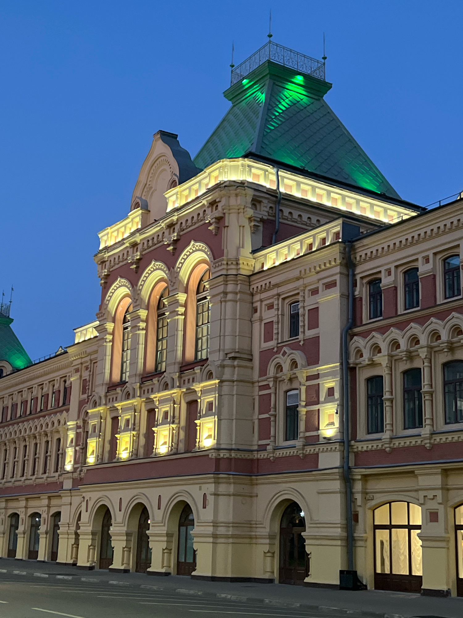 The image shows a side view of a large, ornate building at dusk in Nizhny Novgorod, Russia. The building has a red and cream-colored facade with intricate architectural details, including arched windows and decorative molding. The roof is green and illuminated with bright green lights. The sky is a deep blue, indicating twilight. The building appears to be a historical landmark, possibly a public building or a market. There are no people visible in the image. The street in front of the building is visible, with faint white lines marking the lanes.