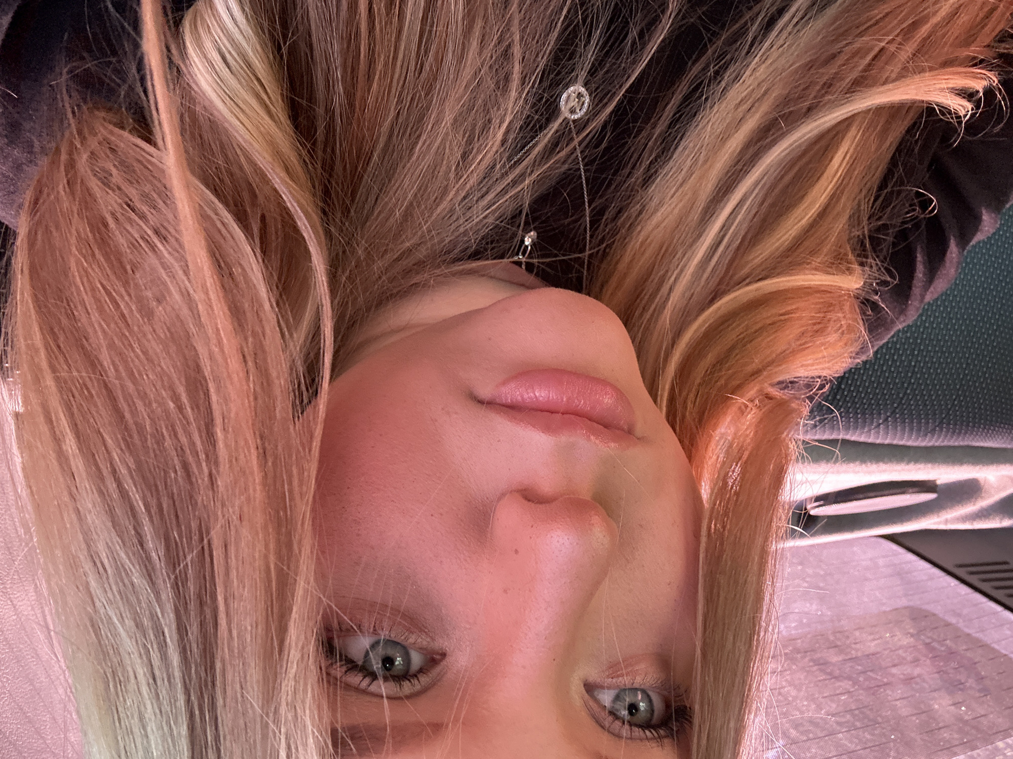A close-up, upside-down portrait of a fair-skinned young woman with blonde hair and blue eyes is visible. She is wearing a necklace with a circular pendant. The background is dark and indistinct. The image does not provide any information about the location of Noviy, Russia.