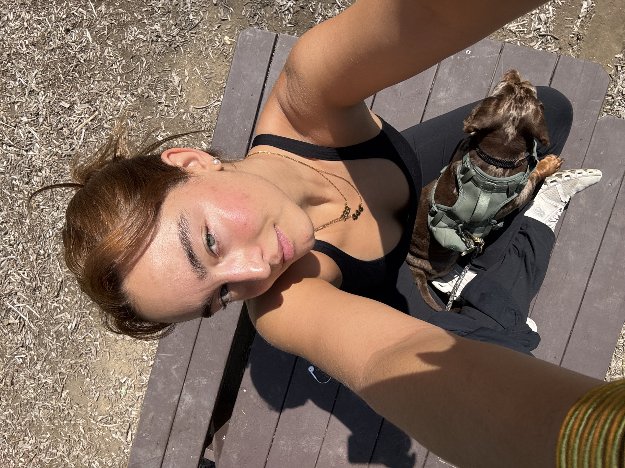 A selfie shows a woman sitting cross-legged on a picnic table with a brown dog in a harness in her lap. The woman wears a black tank top and leggings, along with a gold necklace. She has short brown hair and her left ear has a pearl earring. Her left arm is held out to take the picture and on that wrist there are several gold and green bracelets. The dog is brown and has black ears, and the harness is green. The woman's right foot is visible; she wears white sneakers with black detailing. An earbud can be seen on the table to the right of her arm. The background is a sunny park with leaf litter on the ground. Given the foliage, it is possible that the scene is located in a public park in Los Angeles, United States of America.