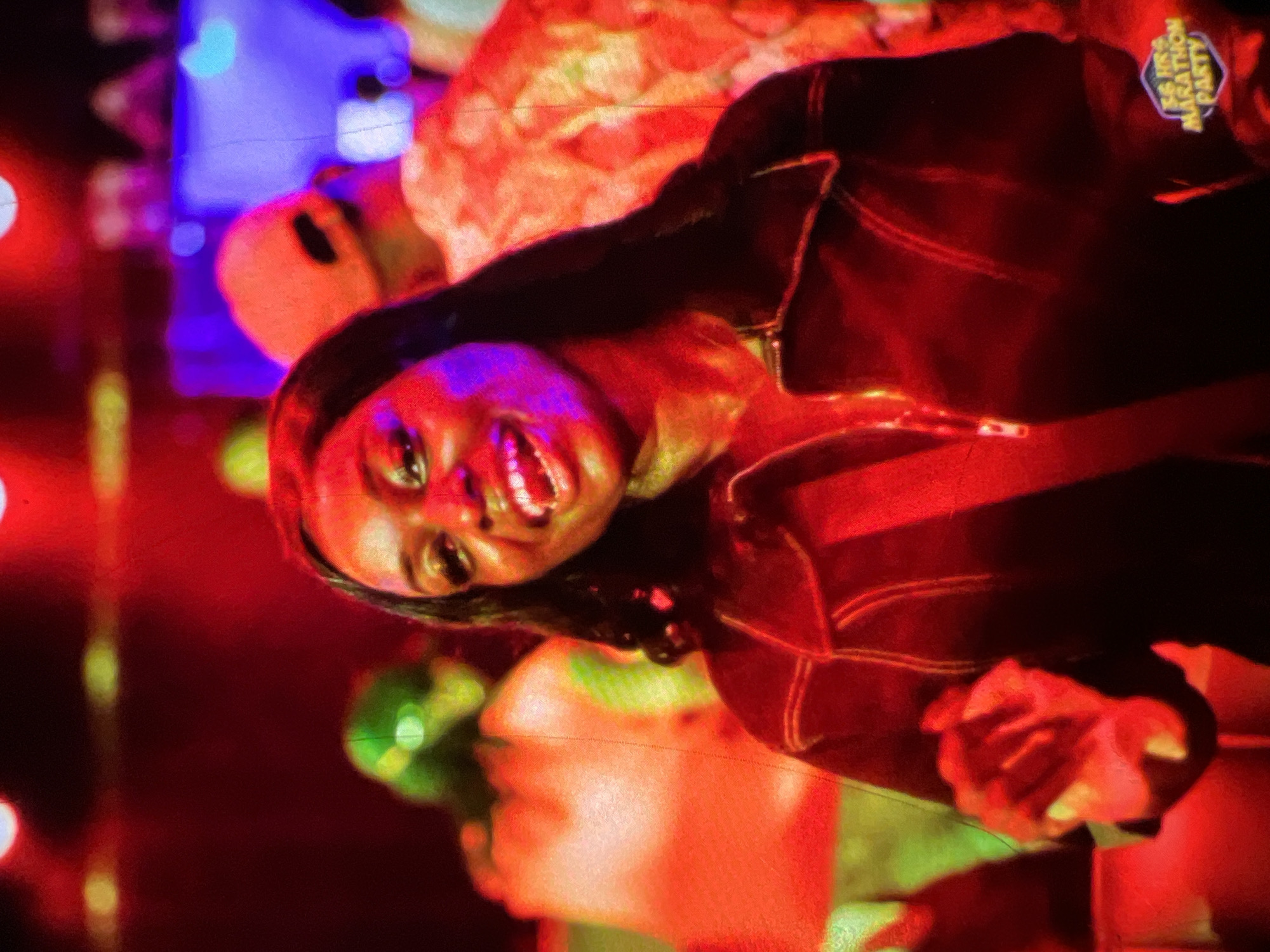 The image is a medium shot of a Black woman smiling broadly with her mouth open and teeth visible. She has long, dark, wavy hair and is wearing a dark jacket with a zipper and contrast stitching. The background is blurred and illuminated by vibrant red and purple lights, suggesting a nighttime event or party. Several other figures are visible in the background, out of focus. To the lower right, a hexagonal badge with the words "72 HRS MARATHON PARTY" is visible. The scene appears to be at an indoor event in Lagos, Nigeria, given the cultural context and the text on the badge.