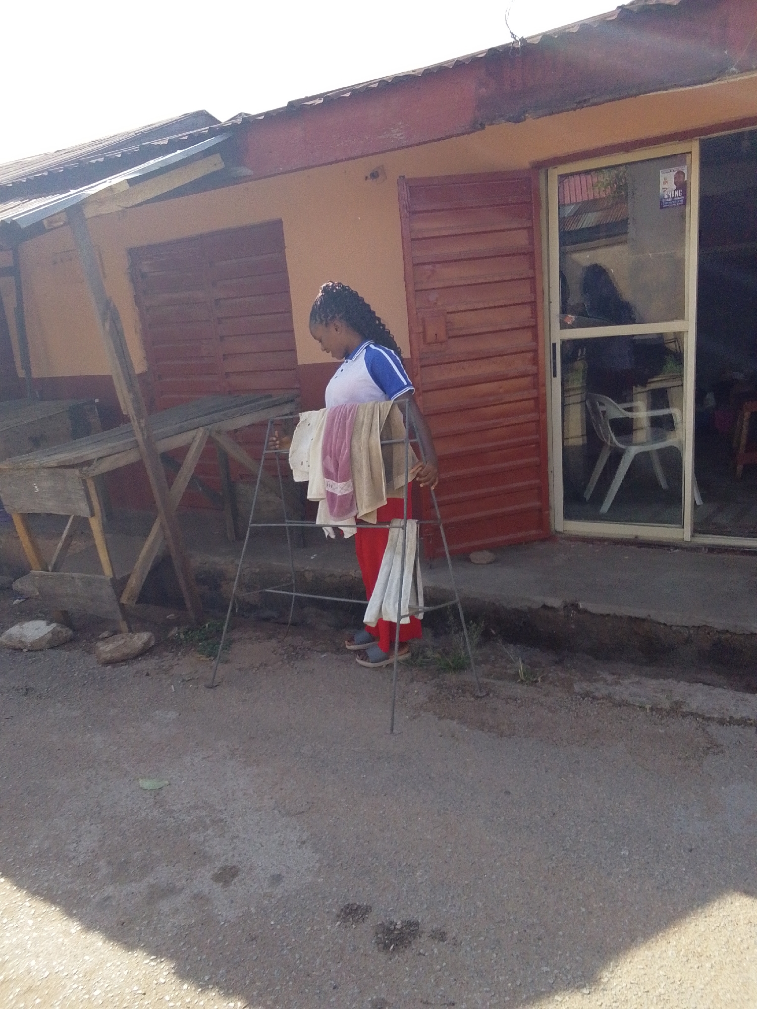 The image depicts a woman standing on a paved road in front of a building. She is holding a metal clothes drying rack with laundry hanging on it. The building has an orange facade with brown trim and a brown wooden door next to a sliding glass door. There is a small wooden table on the left side of the building. The woman is wearing a white and blue shirt, red pants, and gray slippers. She has her hair in braids. The location may be Jos, Nigeria.