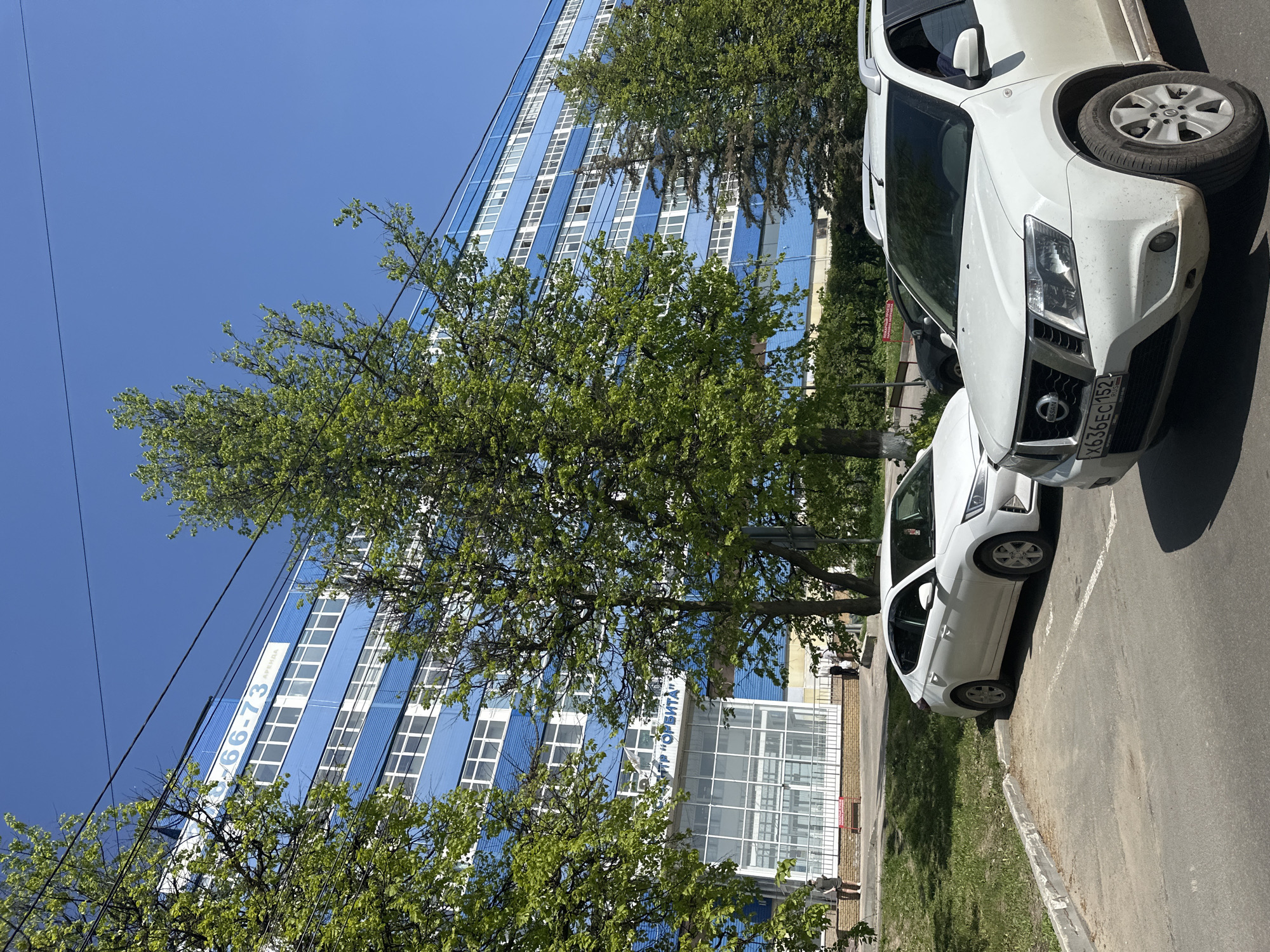 The image depicts a street scene in Nizhny Novgorod, Russia. A tall, blue and white building with numerous windows dominates the background, partially obscured by leafy green trees. A sign on the building reads "ЦТР "ОРБИТА"" which translates to "CTR "ORBITA"" and "АРЕНДА" which translates to "FOR RENT" with a phone number.
Two white cars are parked on the right side of the frame. The front car is partially out of frame, showing the front end and a tire, while the second car is fully visible. The curb and a patch of green grass are visible along the bottom edge of the image. The sky is clear and blue. A black wire crosses the top-left portion of the scene. People are walking near the front of the building. The license plate of the car at the top right of the frame can be clearly seen. The license plate is X636EC 152.