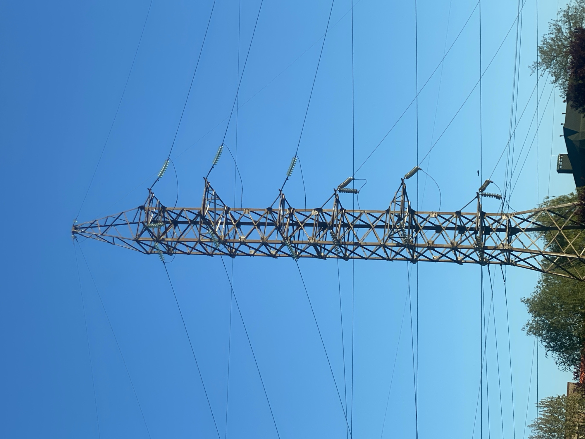 The primary subject of the image is a large, brown metal electricity transmission tower, commonly known as a pylon. The pylon, featuring a complex lattice-work structure of beams, spans horizontally across the frame due to the image's orientation.
Numerous dark, thin power lines extend from the pylon, creating a network of parallel and intersecting lines against a backdrop of a clear, vibrant blue sky. Attached to the pylon are multiple light-colored insulators, appearing to be made of glass or ceramic, which support these power lines. These insulators are visible in various segments along the tower's length, appearing light green or yellowish.
On the right side of the image, parts of green trees and the corner of what appears to be a building are visible, suggesting the pylon is located in an area with vegetation and some structures. The strong, clear blue of the sky indicates bright, sunny weather during the daytime. The photograph is taken from a low angle, looking upwards at the infrastructure. There are no people or specific activities depicted. No text is visible.