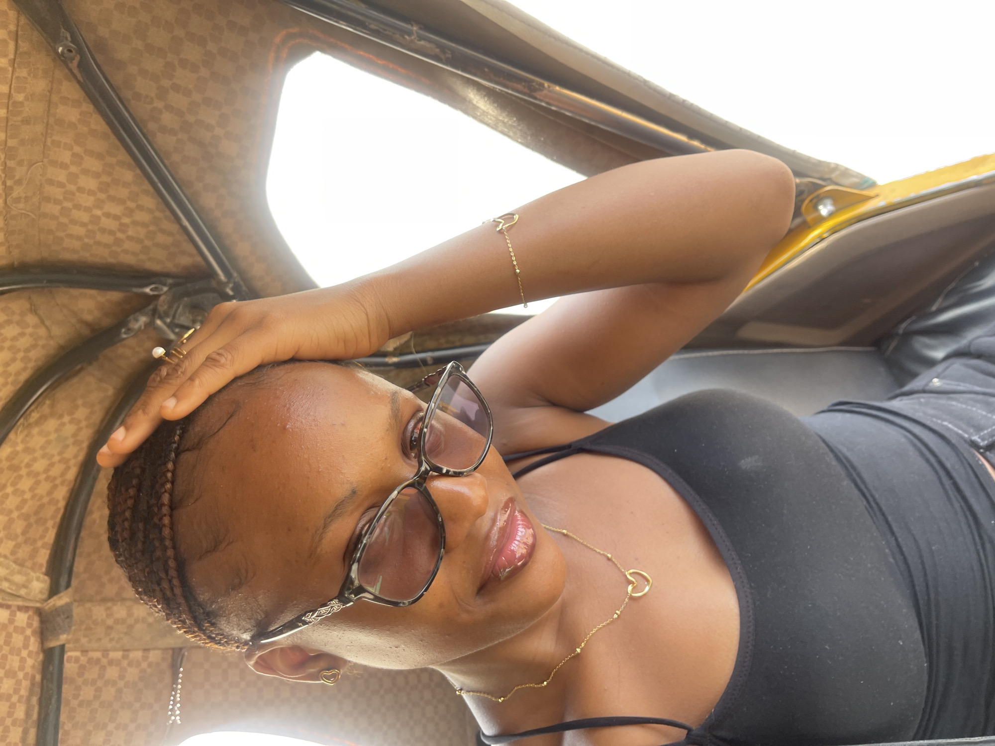 A young Black woman wearing glasses and a black tank top is inside a vehicle, possibly a tuk-tuk, which appears to be a common mode of transportation in Rafin Par, Nigeria. Her hair is braided, and she is wearing a necklace with a heart pendant and a bracelet on her right arm. She has her hand on her forehead, looking downwards, with a slight smile. The interior of the vehicle is visible, with a patterned brown fabric covering the roof and yellow metal framing. The overall lighting suggests it is daytime.