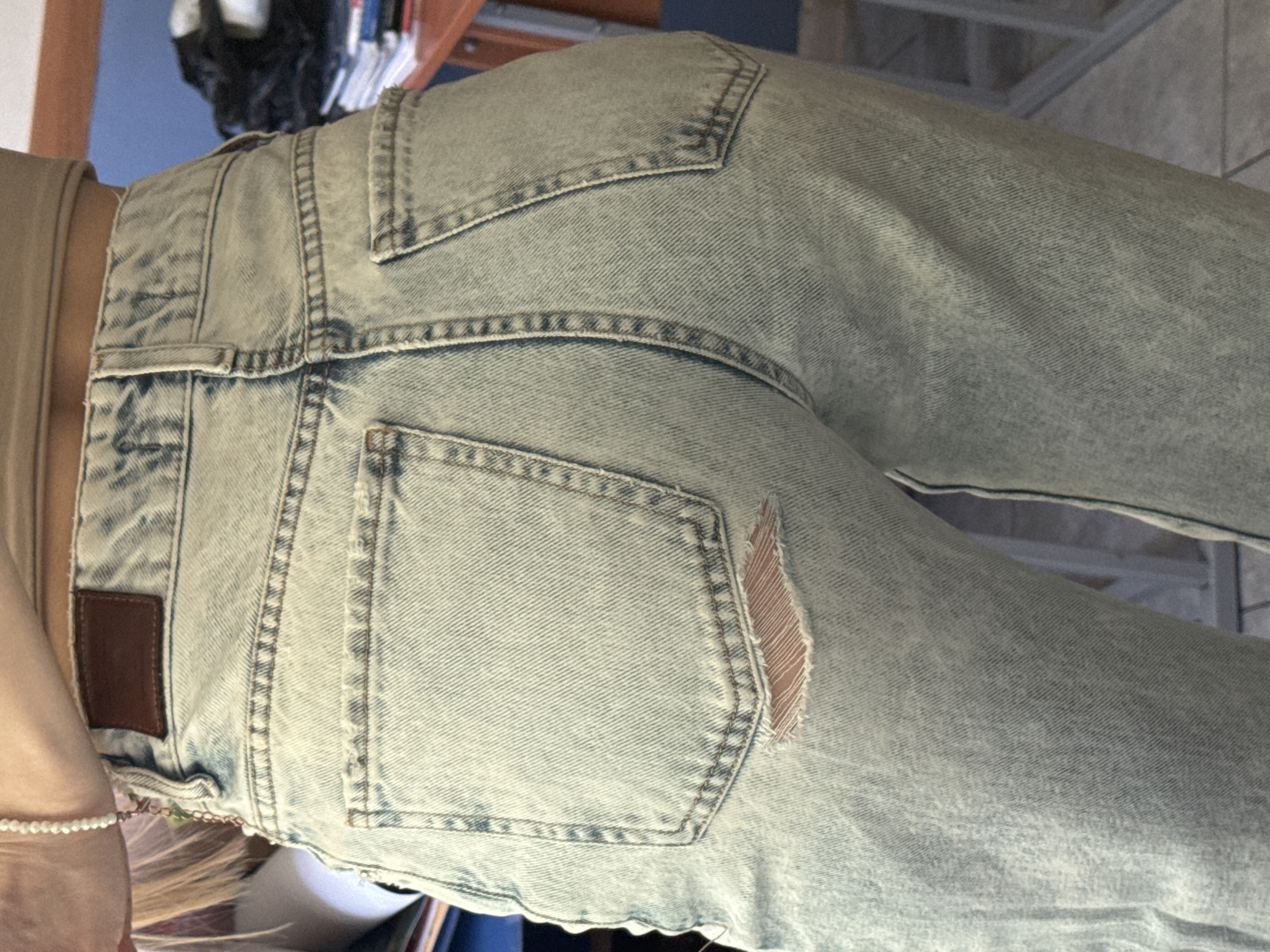 The image shows a close-up of the rear of a pair of light-wash denim jeans worn by a person. The jeans have a standard five-pocket design with visible stitching and a small tear on the right back pocket. The waistband includes a brown leather patch. A small beaded bracelet is visible on the person's wrist. The location is not discernible.