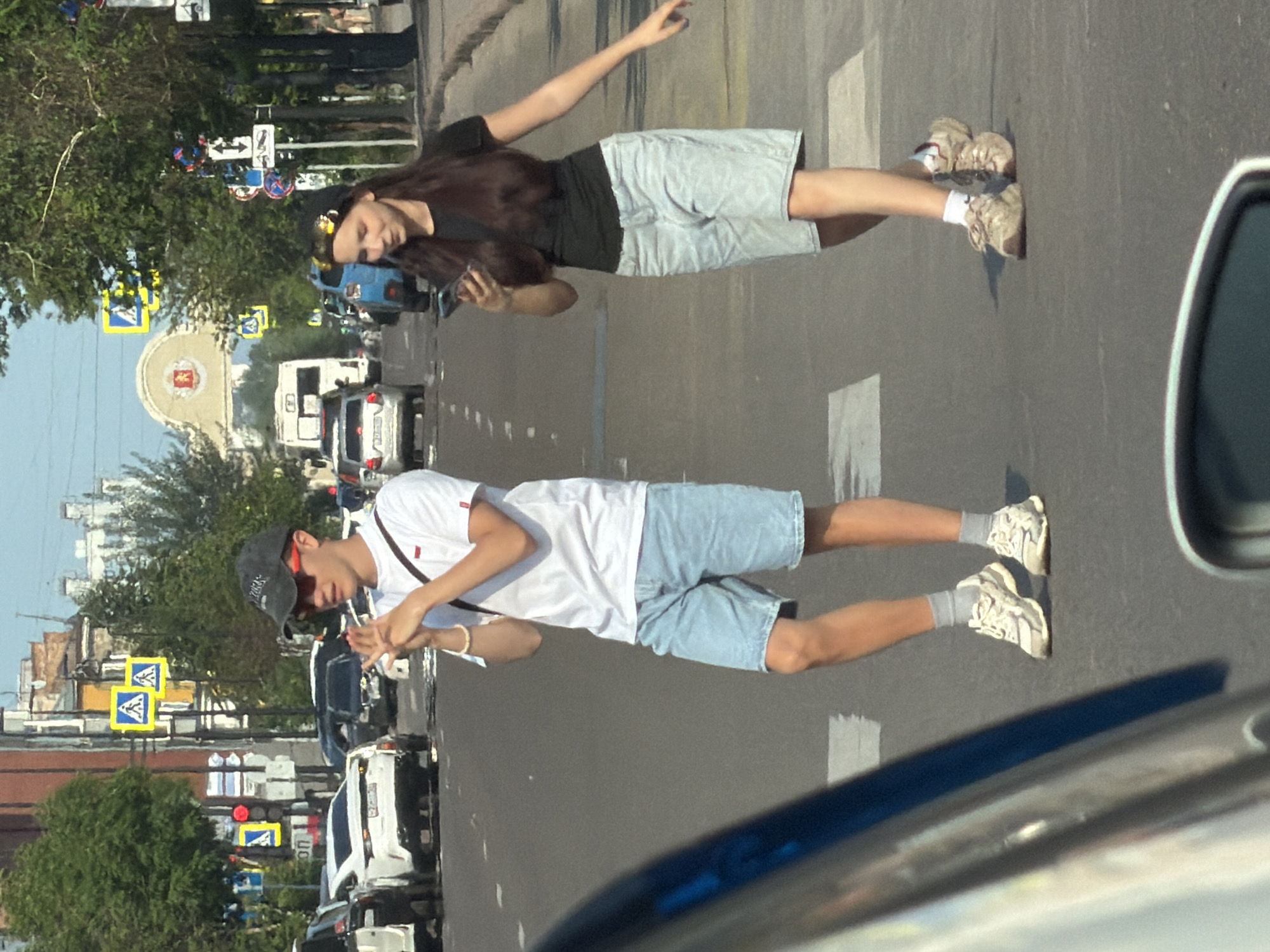The image depicts two people, a woman and a man, on a paved road, likely in Krasnoyarsk, Russia. The woman, in the upper portion of the image, is wearing a black t-shirt, grey shorts, and appears to be lying on the road with her arm extended upwards. She has dark hair and is wearing a beaded headband. A mobile phone is visible in her hand. The man, in the lower portion, is wearing a white t-shirt, blue denim shorts, and a dark baseball cap. He is also lying on the road, facing away from the camera, and his hands are raised. Both individuals are dressed for warm weather.
The scene appears to be a street with buildings and trees visible in the background. Several cars are present, including a white van and a dark-colored vehicle. Traffic signs and lights are also visible, suggesting an urban or suburban environment. The time of day appears to be daytime, with sunlight casting shadows on the road. The overall impression is that of a casual or possibly staged event, with the individuals engaging in an unusual activity on a public road. No discernible text is visible on clothing or objects.