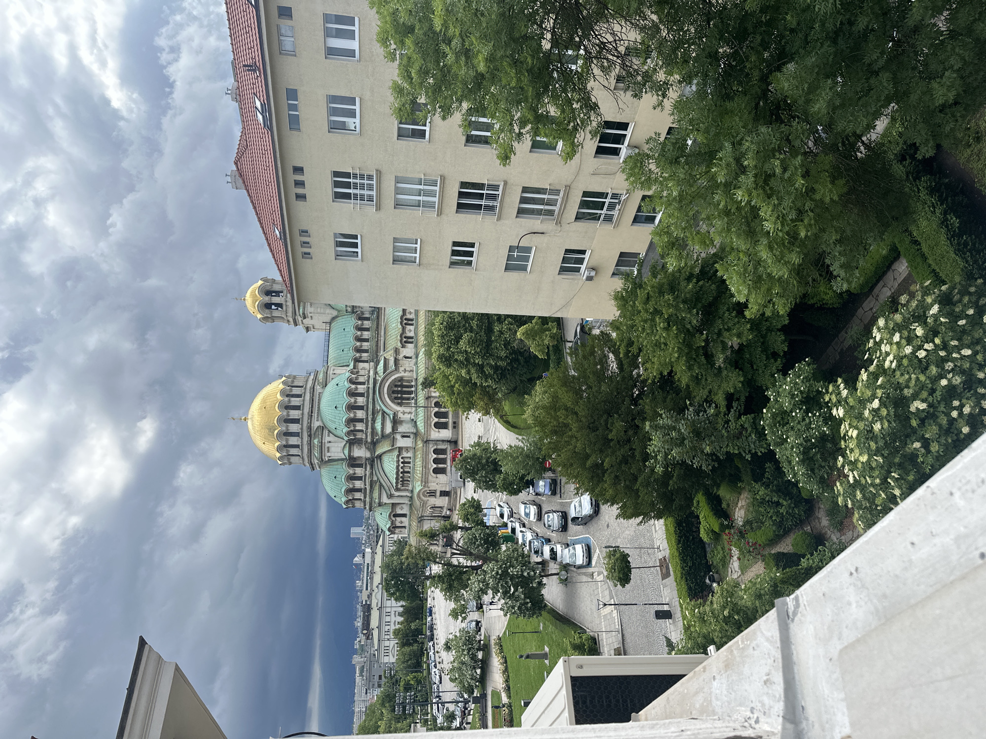 The image captures an urban scene from a high vantage point, looking down and across at a prominent cathedral and surrounding architecture under an overcast sky. In the midground, a grand, ornate cathedral with striking gold and teal-colored domes stands out, showcasing intricate architectural details.
To the right of the cathedral, a multi-story building with a light beige facade and a red tiled roof dominates the right side of the frame. Its numerous windows, some with grilles, are clearly visible. The area between this building and the cathedral, as well as the immediate foreground, is rich with lush green foliage, including tall trees, dense bushes, and flowering plants with white blossoms, indicating a park or garden setting.
Below, a street features several parked cars, predominantly light-colored sedans and SUVs, along a paved or cobblestone surface. Other buildings and urban infrastructure stretch into the distance. The sky is heavily clouded with dark, dramatic grey clouds, suggesting an overcast day with potentially impending rain. A part of an air conditioning unit is visible in the lower right corner, indicating the photograph was taken from a building. No discernible human activity or legible text is visible.