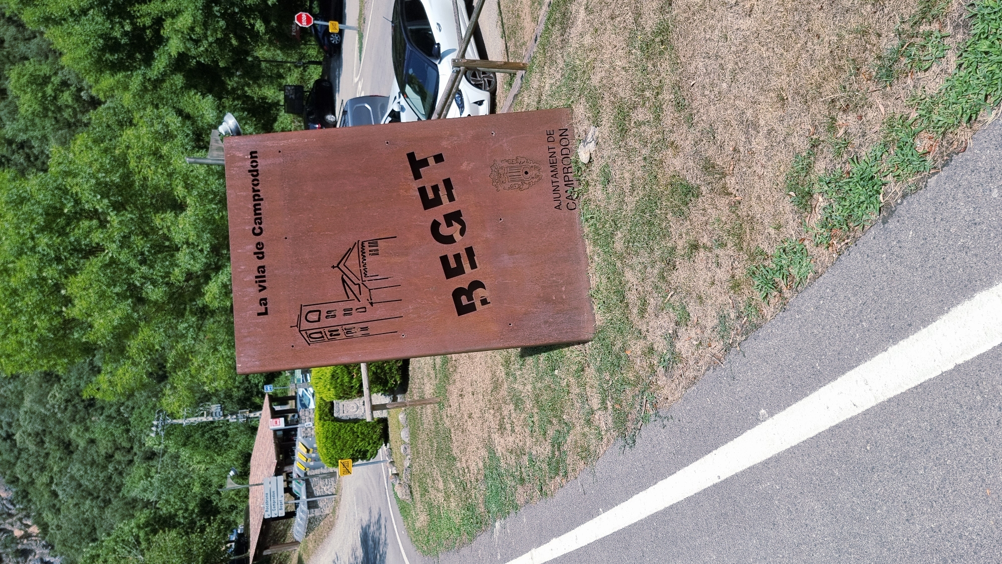 The image shows a large, rusty metal sign with the words "La vila de Camprodon" and "BEGET" cut out. A drawing of a church is also depicted on the sign. Below the word "BEGET" is a coat of arms and the text "AJUNTAMENT DE CAMPRODON". The sign is leaning slightly and is positioned on a grassy area next to a paved road with a white line. In the background, there are trees, a building with a red roof, some parked cars, and a STOP sign. The scene appears to be outdoors in a rural or semi-rural setting.