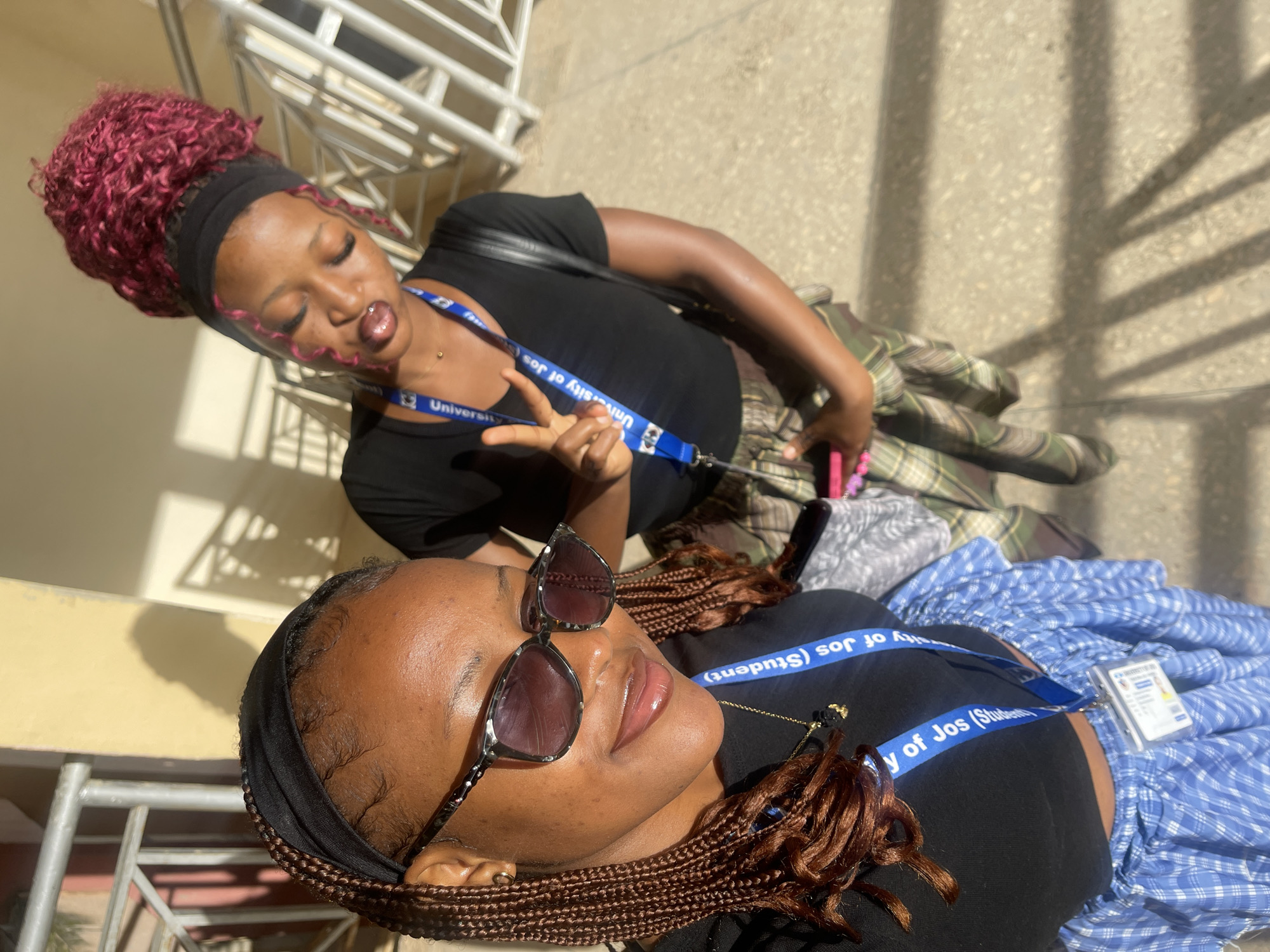 Two young Black women are standing on an outdoor walkway in bright sunlight, casting long shadows. The woman in the foreground is wearing a black headband, a black crop top, sunglasses, and a blue and white checkered skirt. Her hair is braided and falls over her shoulders. She is looking towards the camera and smiling, with her right hand extended towards the camera. A blue lanyard with the text "University of Jos (Student)" is visible around her neck.
The woman in the background is wearing a black headband and a black t-shirt. Her hair is styled in a vibrant reddish-pink bun. She is looking to her left and making a peace sign with her right hand. She is also wearing a similar blue lanyard. Her outfit includes a plaid skirt.
The walkway appears to be paved and there are metal railings and a beige-colored building in the background. The overall scene suggests a casual outdoor setting on a university campus.