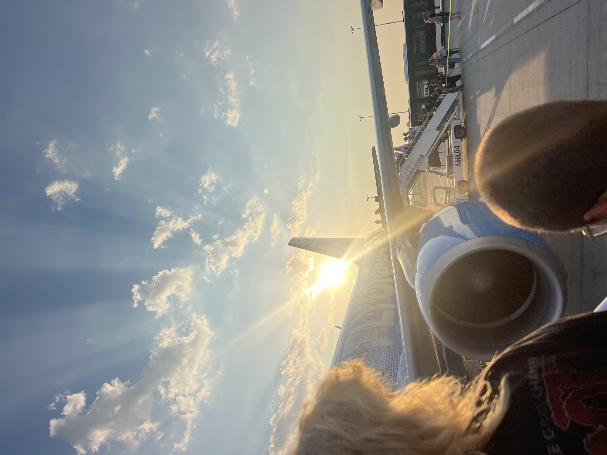 The image depicts an outdoor scene at an airport during sunset. A commercial airplane, primarily blue and white with hexagonal patterned accents and visible text reading "FLYON," is parked on the tarmac. The sun is low in the sky, casting long rays of light through the clouds, creating a dramatic effect. Passengers are boarding the aircraft via a mobile staircase labeled "Aeroport Handling." In the foreground, the backs of two individuals are visible, one with blond, curly hair and the other with short, dark hair, obscuring their faces. Another person is partially visible in the lower right corner. A bus is parked behind the plane, and airport lighting fixtures are present in the background. The overall environment suggests an active airport environment during a sunset, likely at Chișinău International Airport (KIV) in Chișinău, Moldova, given the context of the prompt.