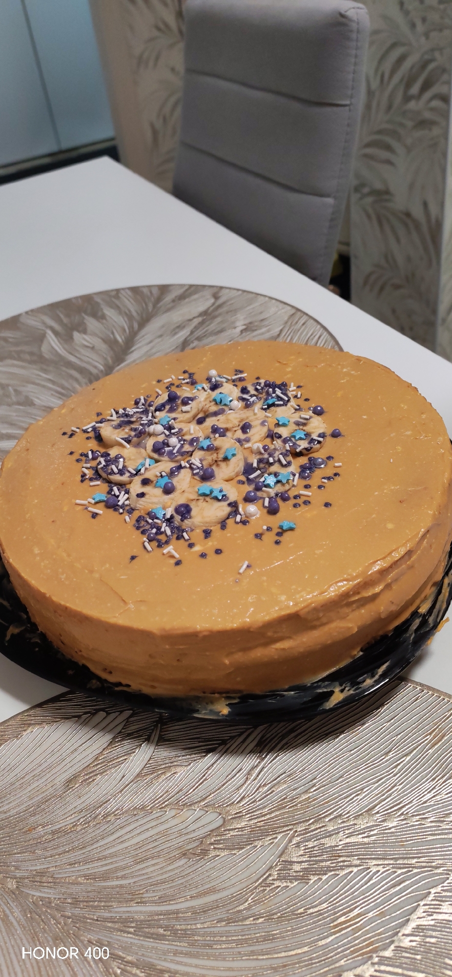 A top-down shot shows a round, caramel-frosted cake resting on a decorative black plate and a table with a gray and white patterned placemat. The cake is decorated with banana slices, purple and white sprinkles, and blue star-shaped candies. In the background, a white table and a gray chair with a ribbed back are visible. The image also has a watermark indicating "HONOR 400" on the bottom.