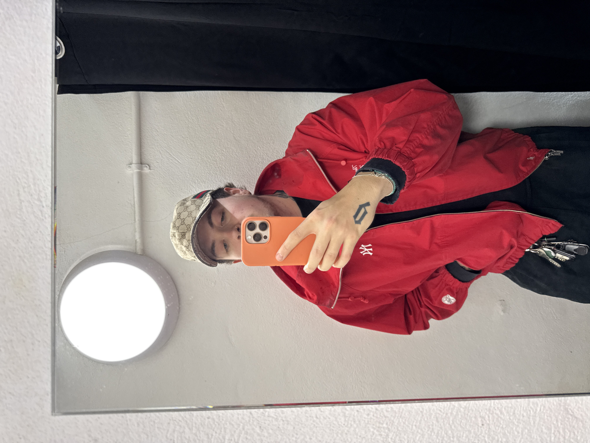 A person wearing a Gucci cap and a red jacket with the New York Yankees logo is taking a selfie in a mirror. The person is holding an orange iPhone. Behind the person in the mirror are a white wall, a circular light fixture, and a black curtain or backdrop. There is no specific information in the image to indicate the location is in London, United Kingdom.