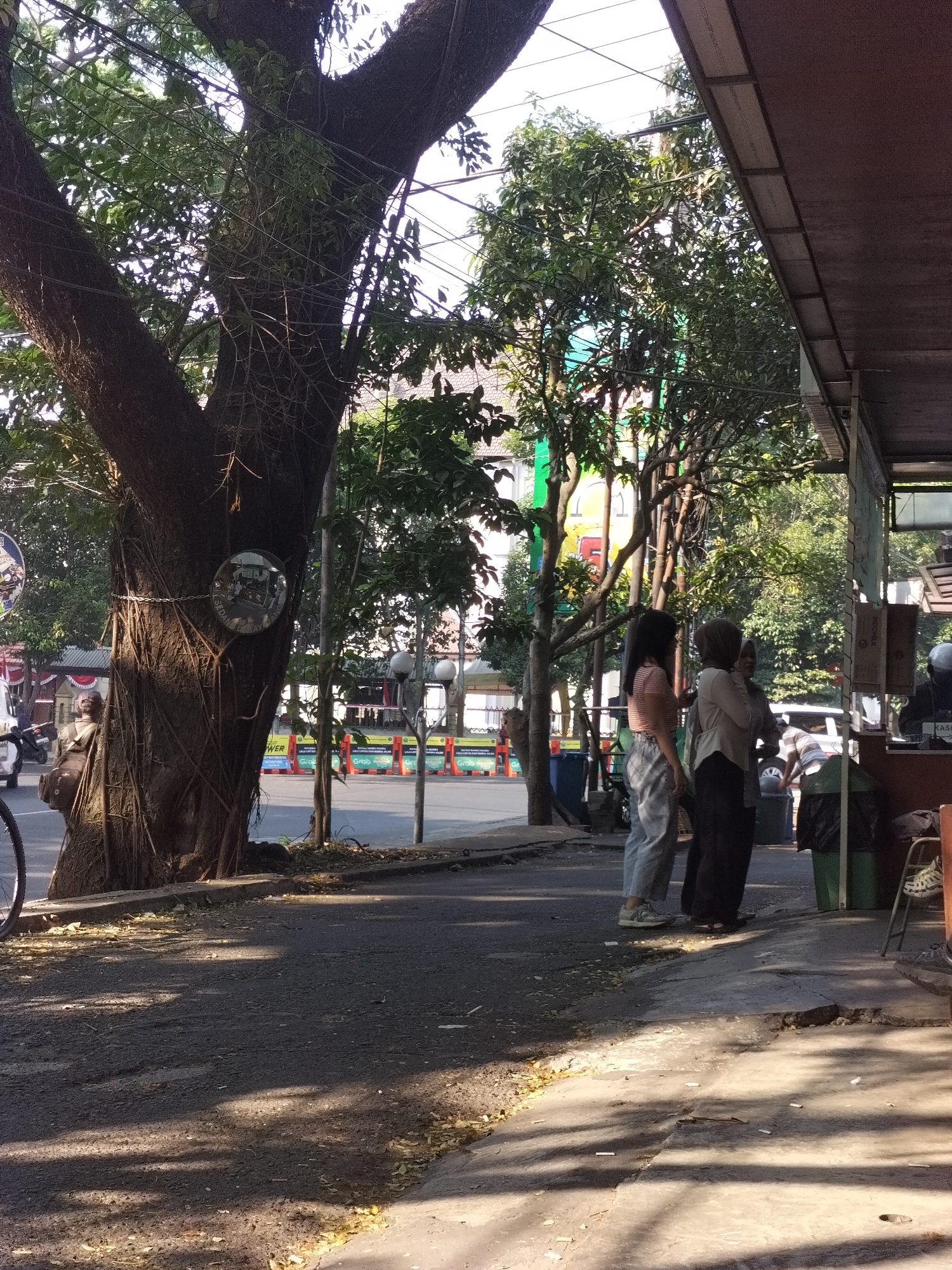 The image depicts a street scene in Malang, Indonesia, with a focus on a large banyan tree on the left. The tree has exposed aerial roots and a circular mirror is attached to its trunk, reflecting the surrounding environment. Two women are standing near a structure on the right side of the image, engaged in conversation. One woman is wearing a light-colored hijab and a long-sleeved top, while the other has dark hair and is wearing a striped t-shirt and light-colored pants. A bicycle is partially visible on the far left. The street in front of them has patches of sunlight and shadow, with scattered leaves and cigarette butts on the pavement. In the background, there are trees, buildings, and traffic barriers. The overall atmosphere is that of a casual urban setting on a sunny day.