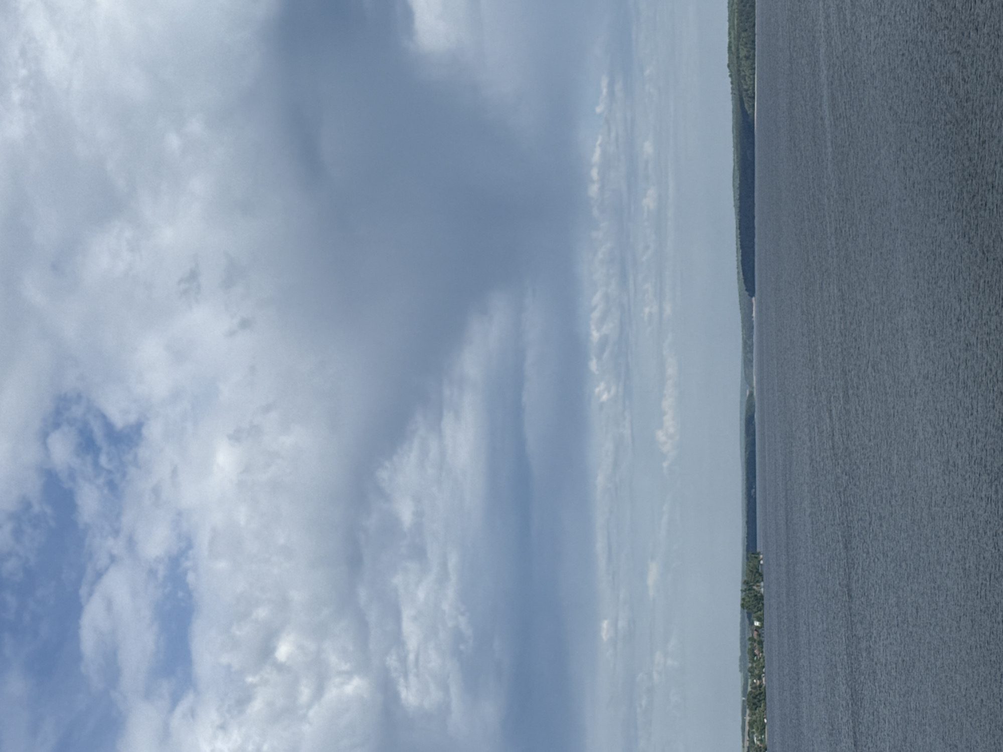The image depicts a view of a body of water, likely a reservoir or large lake, with a forested shoreline in the city of Votkinsk, Russia. The sky is predominantly filled with clouds, ranging from light and scattered to dark and dense. The water appears calm with slight ripples. The shoreline is densely wooded. There is no evidence of people visible.