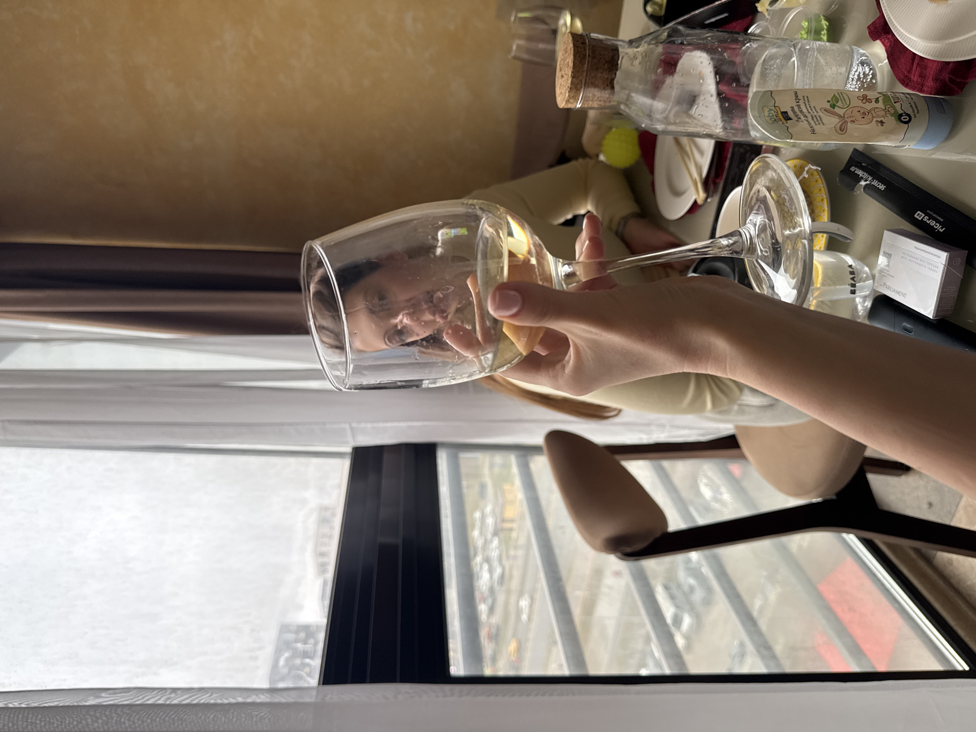 The image shows a person holding a wine glass in front of a window. The window offers a view of a city, which is likely in Unknown, Unknown. The person is reflected in the glass. In the background, there's a cluttered table with various items, including a cork-topped bottle, plates, cutlery, and a container of baby cream. The room is illuminated by natural light coming through the window. A chair is visible near the window.