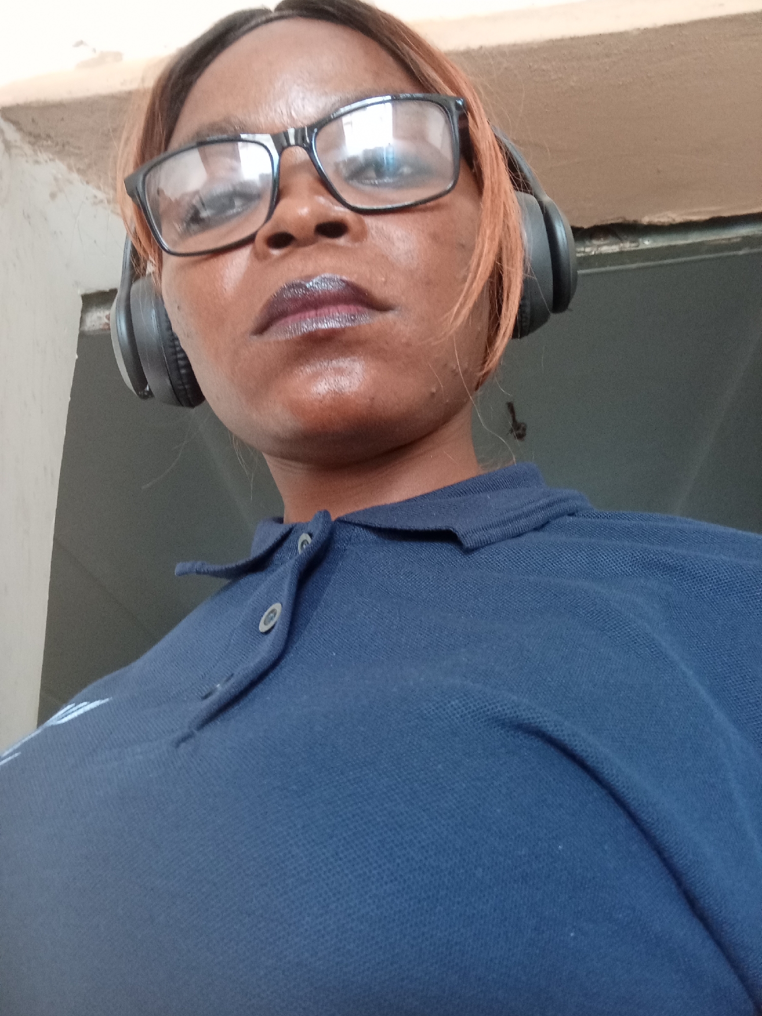 A woman with dark skin, light brown hair, wearing black-rimmed glasses and black headphones, looks directly at the camera. She is wearing a dark blue polo shirt with two buttons at the collar, one of which is visible. Her lips are painted with dark lipstick. The background consists of a wall and a ceiling. The image appears to be a selfie taken indoors, possibly in Jalingo, Nigeria, given the context provided.