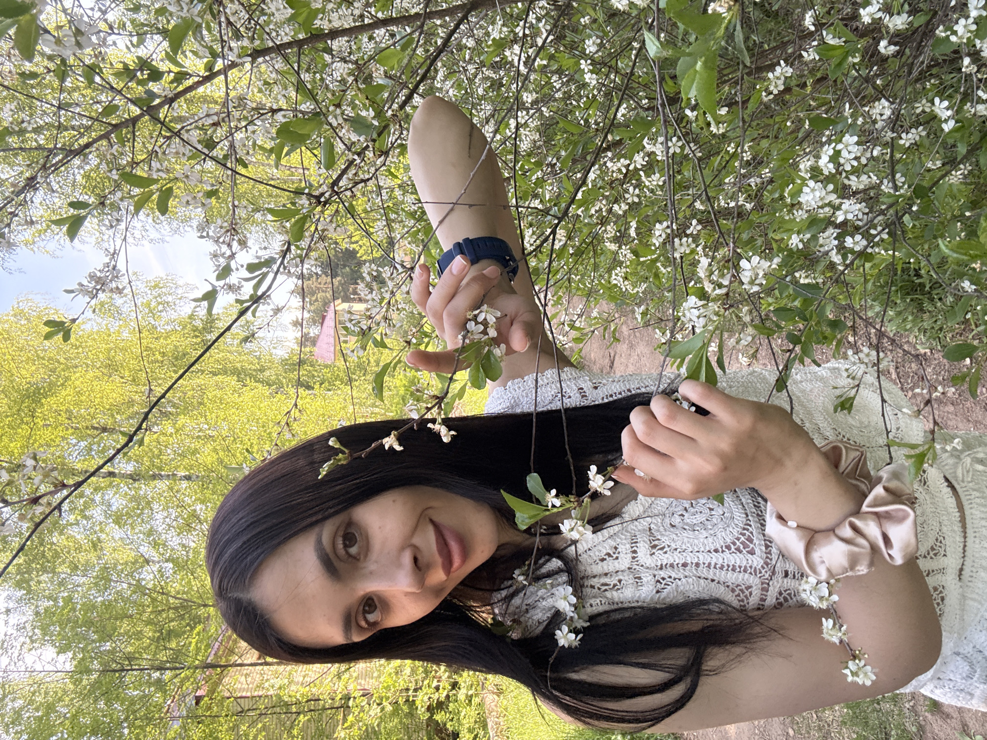 A dark-haired woman is lying beneath a blooming tree, likely a cherry tree based on the white blossoms, in what appears to be a park or outdoor setting. She is wearing a white crocheted top. She has a blue wristband on her right wrist and a tan scrunchie on her left. The background includes foliage suggesting a natural or park area. There is a glimpse of a pink flag or decoration in the distance, though not sharp or clear enough to identify its exact shape or purpose. Given the location, this could potentially be a park in Votkinsk, Russia.