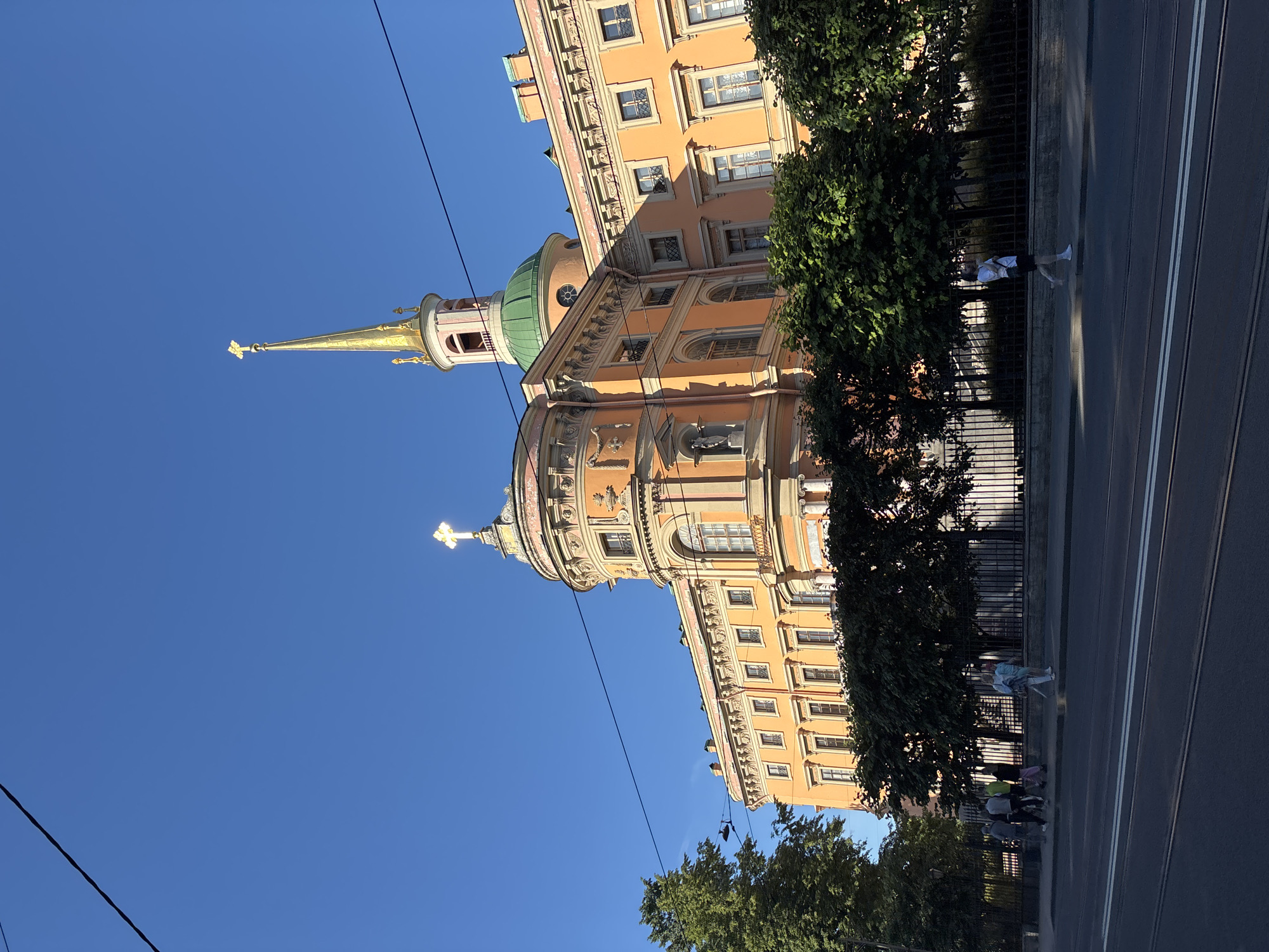 The image shows the Mikhailovsky Castle, also known as the Engineers' Castle, in Saint Petersburg, Russia. The castle is an orange building with a green dome and a tall golden spire topped with a cross. The sky is a clear blue. In the foreground, there is a sidewalk with people walking, and a street with tram tracks. Trees line the sidewalk in front of the castle, casting shadows. The lighting suggests it is daytime, likely in the late afternoon or early evening due to the long shadows.
