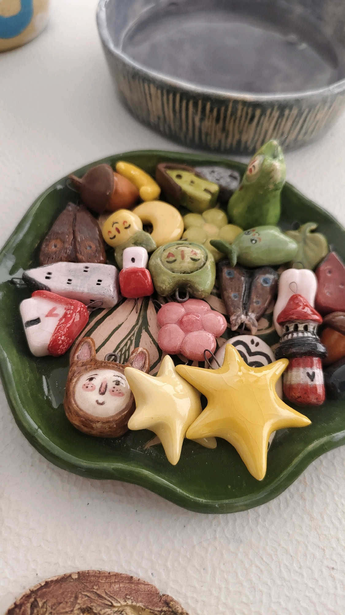 The image shows a collection of small, handcrafted ceramic charms arranged on a green, leaf-shaped ceramic plate. The charms are diverse in shape and color, featuring various objects and figures such as fruits, vegetables, animals, and architectural elements. Among the visible charms are an acorn, a yellow banana-like shape, green avocado halves, a green frog, a white and red rectangular object with markings, a red and white house-like object, a pink flower, a brown animal face with large ears and rosy cheeks, two yellow stars, and a red and white lighthouse. The plate is placed on a white textured surface. In the background, a portion of a large, dark ceramic bowl with a ribbed texture is visible. The scene is well-lit, with natural light casting soft shadows. There are no people or animals present in the image, and no specific location within Krasnoyarsk, Russia can be identified.