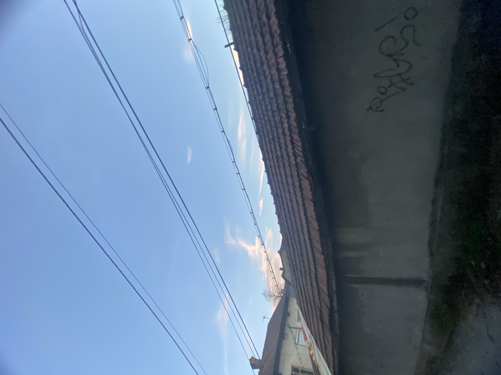 The image shows a view looking up at a building and power lines in Troyan, Bulgaria. A weathered building with a red-tiled roof is visible at an acute angle. A gray concrete structure runs vertically to the right of the building, marked with graffiti. Overhead, several power lines stretch across a blue sky with sparse clouds.