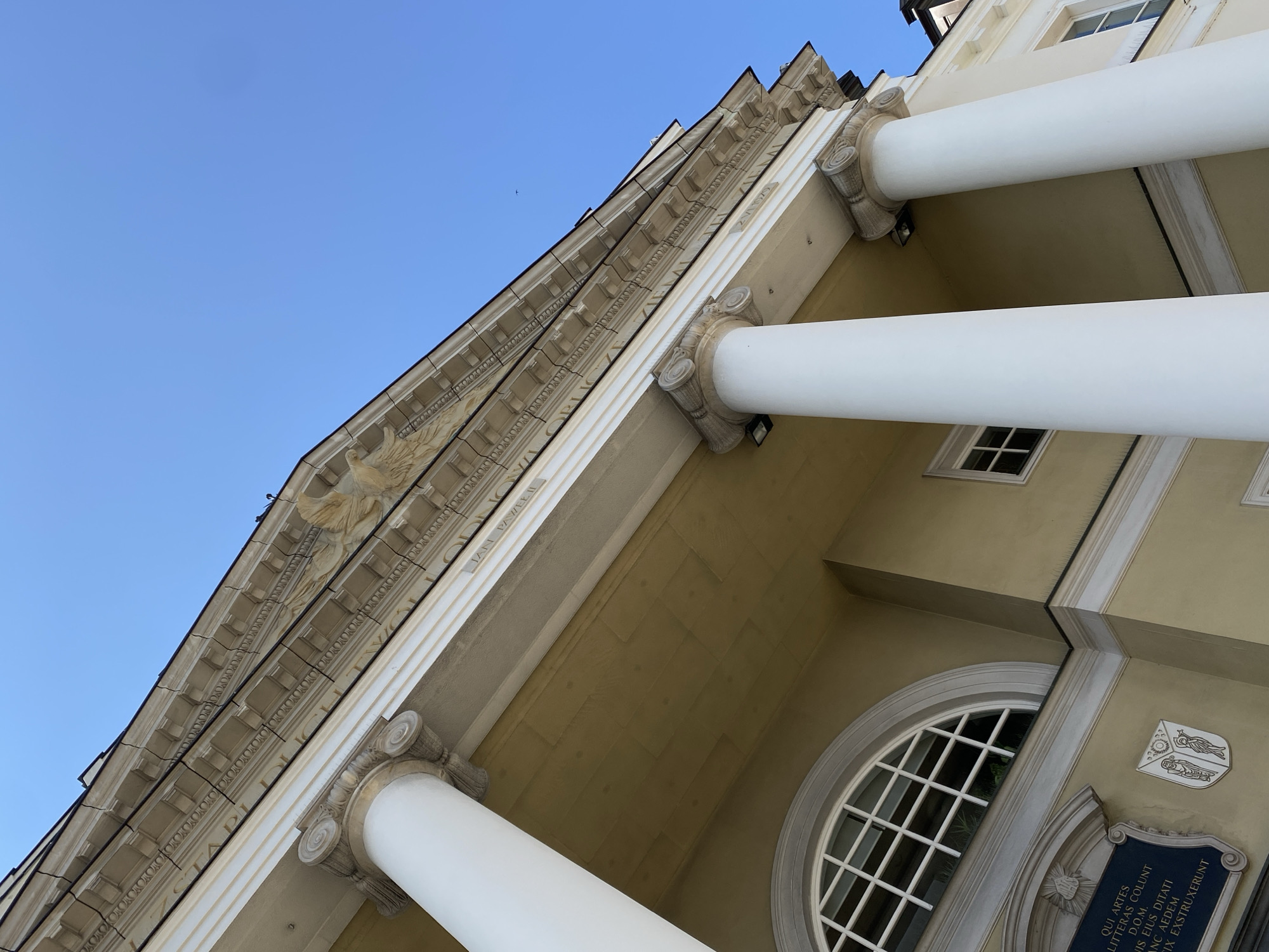 The image shows the facade of a building with classical architectural elements. Three large white columns with Corinthian capitals support an entablature that features decorative moldings and sculpted reliefs. Above the entablature, the building's roofline is visible against a clear blue sky. On the left side of the building, a semi-circular window with a grid pattern is set into a pale yellow wall. Below the window, a dark blue plaque with Latin inscriptions and a coat of arms is mounted on the wall. The architecture suggests a historical or institutional building. This is the building of the Old University of Warsaw, located at Krakowskie Przedmieście 26/28 in Warsaw, Poland.