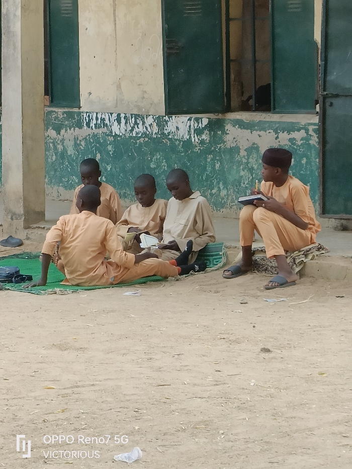 This image captures a group of six young males, likely children or adolescents, engaged in an outdoor learning session in Dutse, Nigeria. All subjects are dressed in similar light-colored (beige or light orange) long robes or tunics and trousers, suggesting a uniform or traditional attire. Most have short, dark hair, while one boy on the far right wears a dark cap.
The central activity involves the boys studying or learning. One boy on the far right sits on a raised step or curb, holding a notebook and pen, seemingly writing or taking notes, and appears to be leading or instructing. The other five boys are seated on the sandy ground, either directly or on green mats. Two boys sit on a solid green mat, while others are on or near a green prayer rug with a geometric pattern. They are looking at books or notebooks, indicating reading or studying. One boy in the foreground sits with his back to the viewer, also seemingly engaged in reading.
The setting appears to be an informal outdoor classroom or study area beside a weathered, old building. The building's walls show peeling paint in green and off-white/yellowish tones, with dark window openings or shutters. The ground is predominantly sandy or dusty earth. Scattered around are a pair of black sandals, a black bag, and a small white plastic bag or piece of paper in the foreground. The bright, diffused daylight suggests it is daytime, possibly morning or afternoon, in dry, warm weather. The boys appear focused and calm, dedicated to their learning.
Visible text in the bottom left corner includes a camera watermark "OPPO Reno7 5G" and "VICTORIOUS".