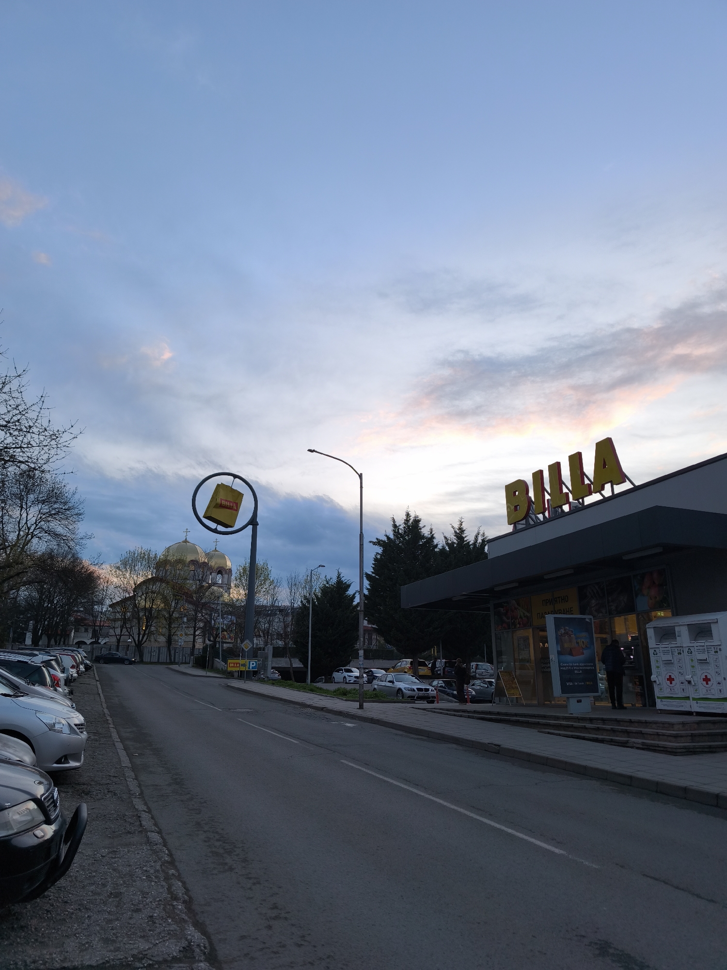 The image captures a street scene in Burgas, Bulgaria, at dusk. Dominating the right side is a Billa supermarket, marked by its prominent "Billa" sign atop the building. Several people are present near the entrance, possibly customers. To the left of the supermarket, a street stretches into the distance, lined with parked cars on the left and some sparse trees. In the background, a distinctive golden-domed building, likely a cathedral, is visible. A Billa advertisement featuring a shopping bag is installed on a street lamp. The sky is a mix of blues and pinks, indicating either sunrise or sunset.