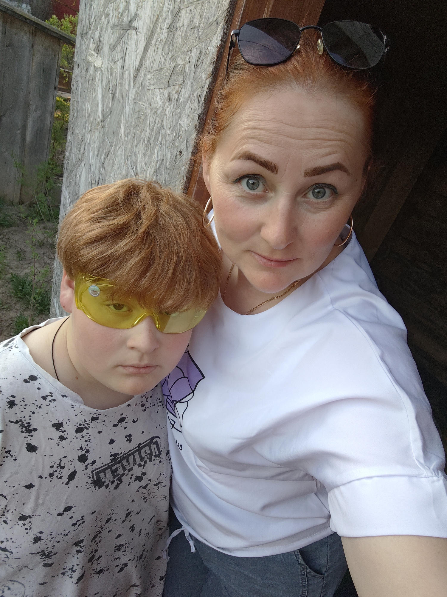 A fair-skinned woman with red hair and sunglasses on her head stands next to a fair-skinned, red-haired child wearing yellow safety glasses. Both are looking at the camera. The woman wears a white shirt and gold hoop earrings and a gold necklace. The child wears a white t-shirt with black splatter marks. The background is a roughly finished plywood wall next to an old gray wooden fence. The location is likely Фоки, Russia, based on the user query.