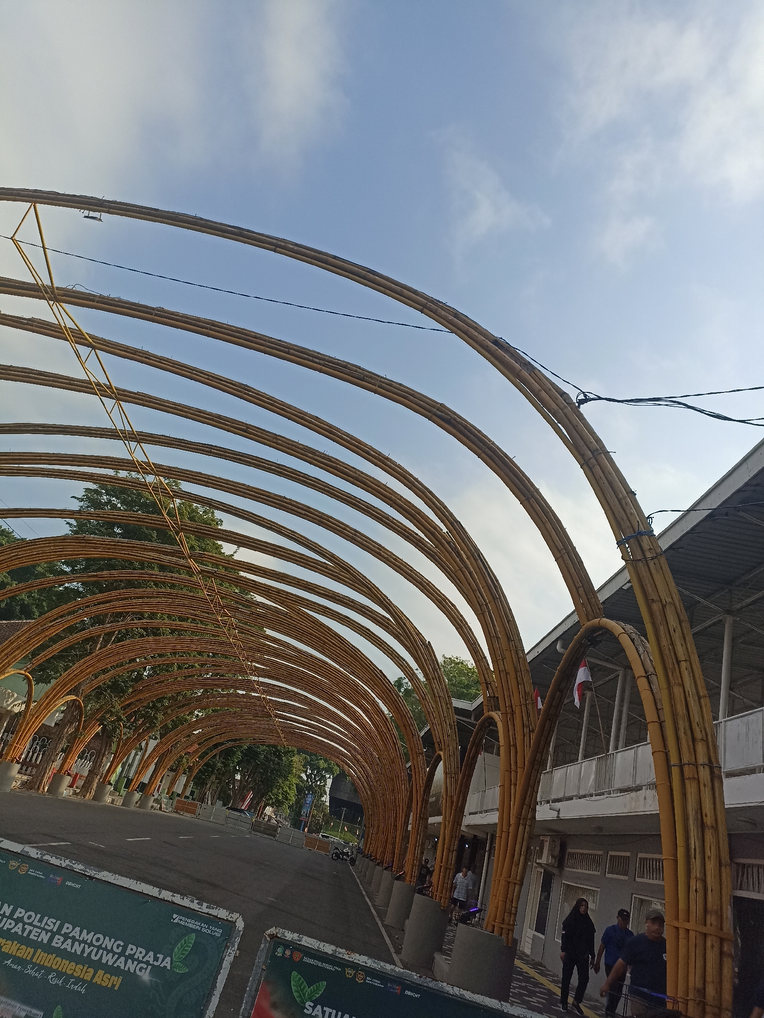 The image shows a street scene in Kelurahan Kepatihan, Indonesia, under a large arched structure made of bamboo. The bamboo structure forms a tunnel-like pathway over the road, with multiple arches extending into the distance. The sky above is partly cloudy and light blue. On the right side of the road, there is a building with a gray facade and white railings on an upper level. An Indonesian flag is visible on the building. Several people are walking on the sidewalk in front of the building. On the left side of the road, there are green banners with text and illustrations. The road itself appears to be a two-lane street with white markings. Trees are visible behind the bamboo structure.