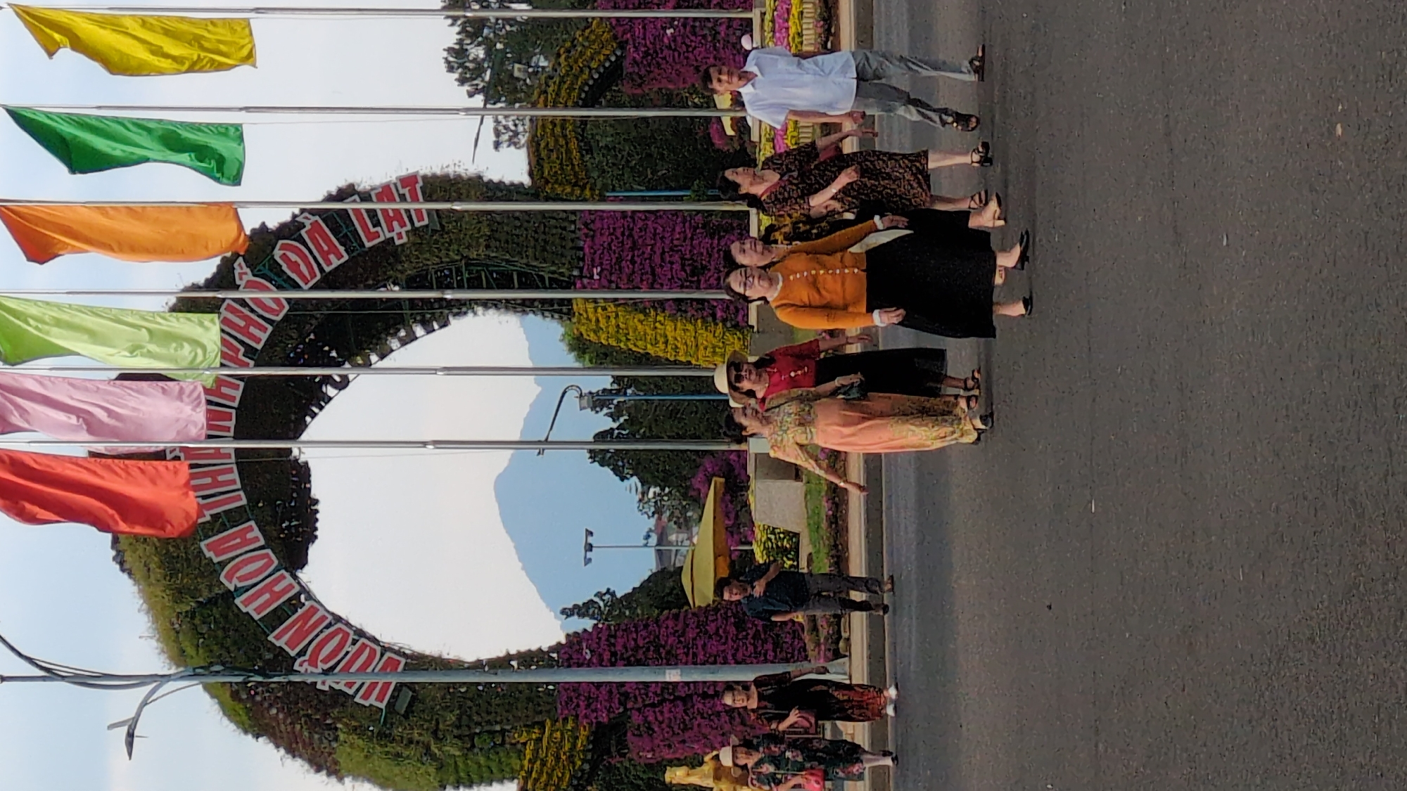 The image shows the roundabout at the entrance to the Da Lat Flower Garden in Da Lat, Vietnam. The roundabout has a large, circular floral display with the words "Vườn Hoa Đà Lạt" (Da Lat Flower Garden) written on it. Above the display, there are several poles with colorful flags. A group of about eight people is standing on the road near the roundabout. The background includes some trees and a distant mountain. The scene appears to be taken during the day in bright sunlight.