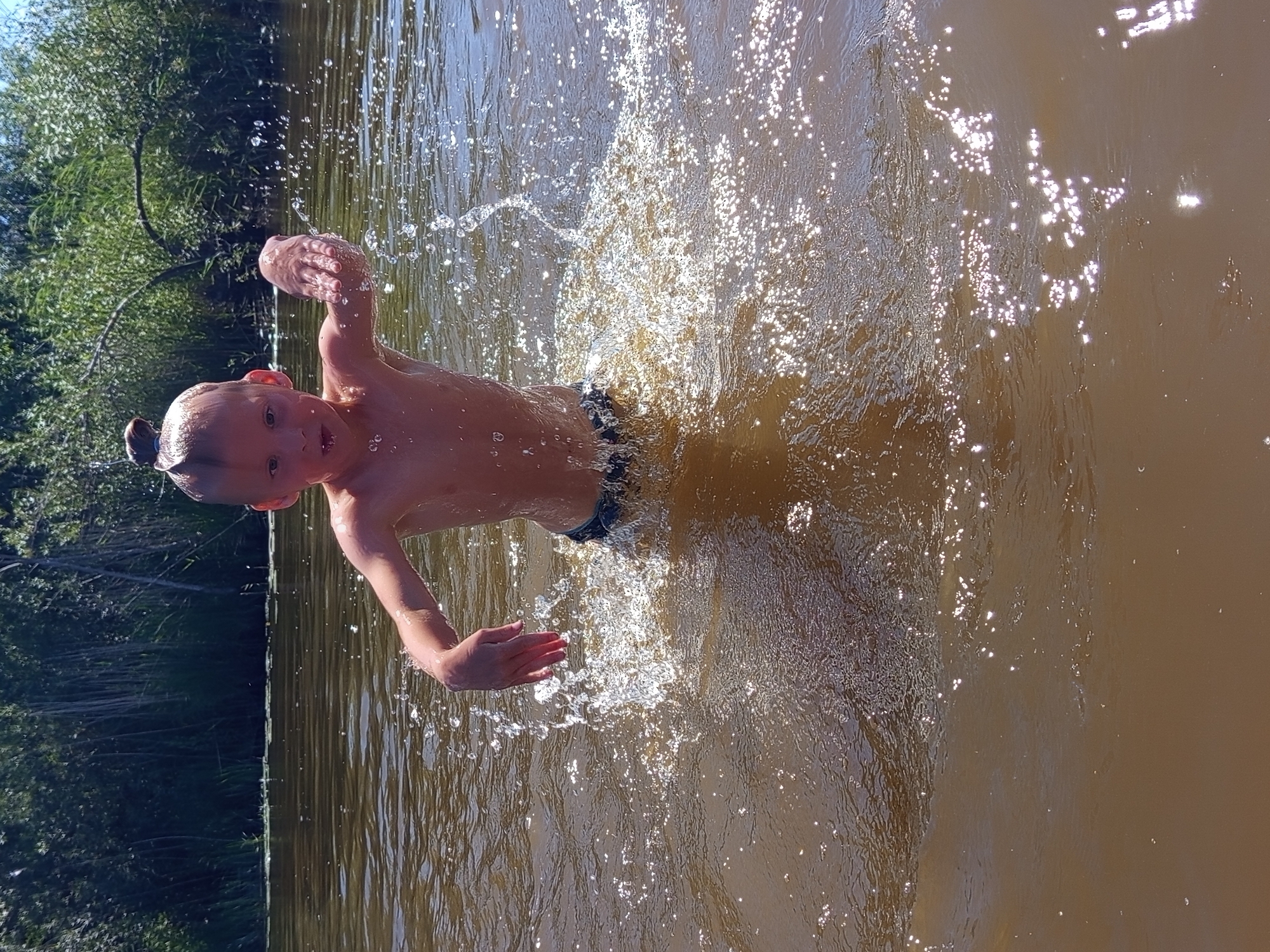 A young child is captured mid-splash in murky brown water, likely a river or lake, in Ostrovtsy, Russia. The child, wearing dark swim trunks, has their arms outstretched and their face turned towards the camera with a surprised or joyful expression. Their hair is slicked back and tied in a small bun. Sunlight glints off the water, creating a sparkling effect around the child. The background features lush green trees and foliage, suggesting a natural, outdoor setting. The time of day appears to be sunny, likely during daylight hours. The overall mood of the image is playful and energetic, highlighting the pure fun of a summer day spent swimming. There is no visible text in the image.