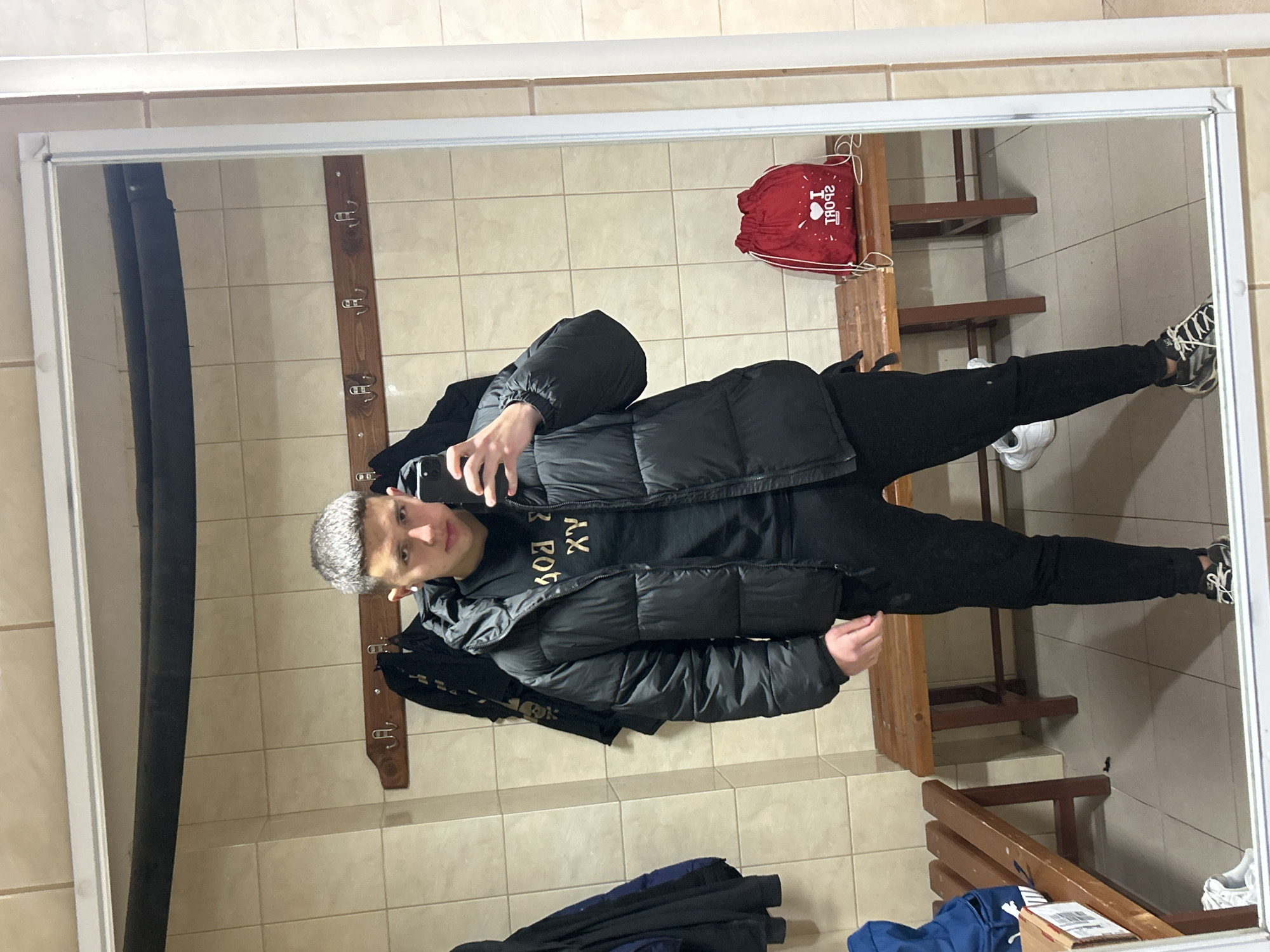 A young male is captured in a mirror selfie, standing upright in what appears to be an indoor changing room or locker room. He has short, light-colored hair and looks directly into the camera with a neutral expression. He is wearing a black puffer jacket over a black t-shirt with gold lettering (appearing as reversed "AX FOR" or similar due to the mirror), black pants, and black and white athletic shoes. He holds a smartphone in his left hand to take the picture.
The reflection in the mirror reveals a room with light-colored, possibly beige or cream, square tiles covering both the walls and the floor. To the subject's right, a tall, vertical wooden plank with several metal coat hooks is mounted on the wall. Below this, and extending behind the subject, are multiple wooden benches. To the subject's left, on one of these wooden benches, rests a red drawstring bag. The bag features a white design and reversed text that reads "I ❤️ SPORT". Outside the bottom frame of the mirror, some dark clothing, including what looks like a blue jacket, is visible on the floor or a lower bench. The setting suggests a functional space, characteristic of a gym or sports facility in Zlatograd, Bulgaria.