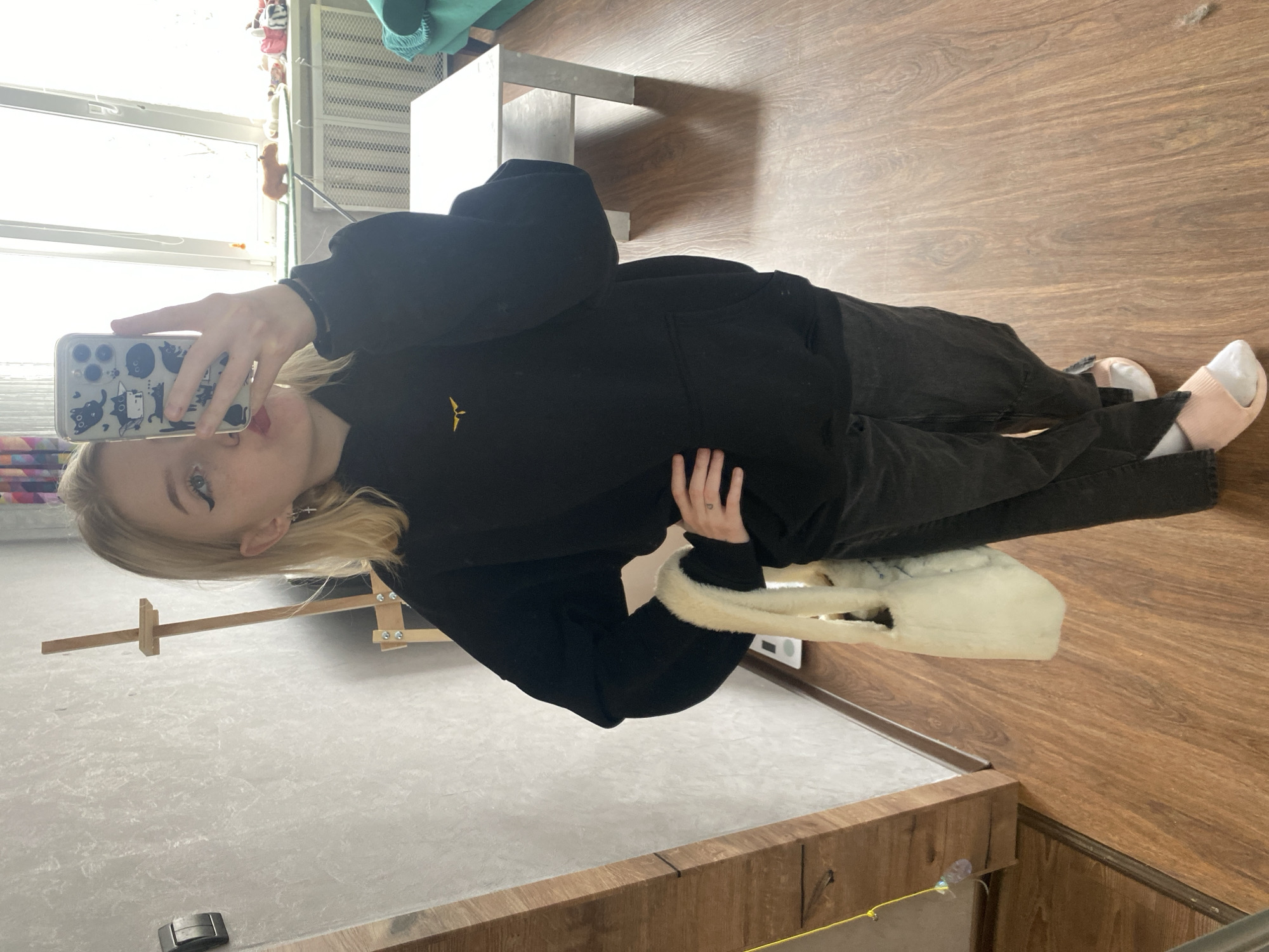 A woman is standing indoors, taking a selfie with her phone. She has blonde hair, is wearing a black sweatshirt, black pants, and pink slippers. She is holding a white purse. Behind her are a window, a table, an easel, and wooden floors. The location is likely in Izhevsk, Russia, but there's not enough detail to confirm this.
